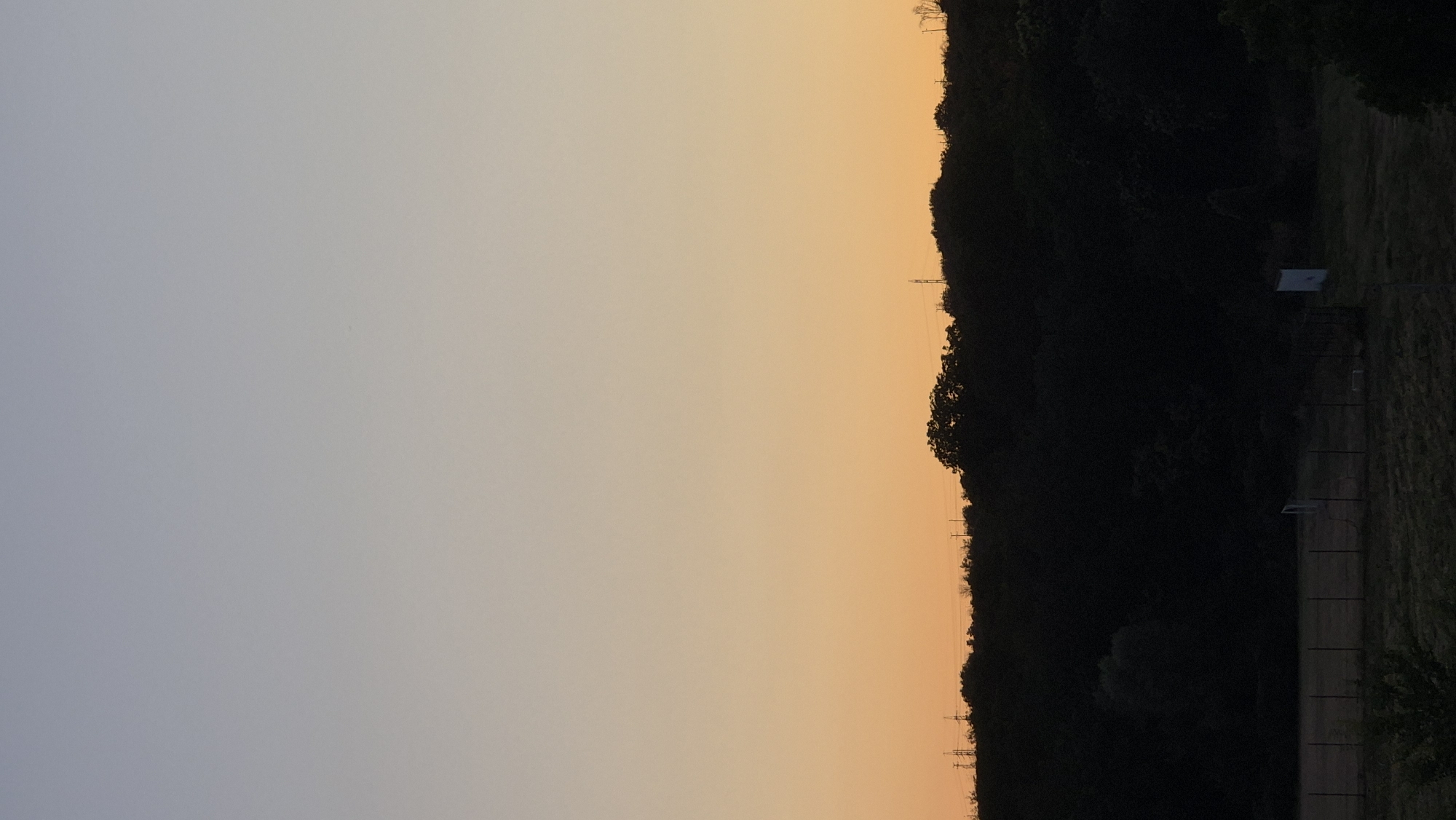 The image depicts a rural or semi-rural landscape at dusk or dawn. The sky transitions from a pale blue at the top to a warm orange hue near the horizon. A dense line of dark trees forms a silhouette against the illuminated sky, suggesting a hill or elevated terrain. Several electricity pylons are visible interspersed among the trees, indicating the presence of power infrastructure. In the foreground, a fenced-off area, possibly a sports field or enclosure, is visible. A small, white rectangular structure stands within this area. The ground appears to be covered in grass or low vegetation, with some shadows cast, indicating the low angle of the sun. There are no people visible in the image. The location is identified as Lovech, Bulgaria.