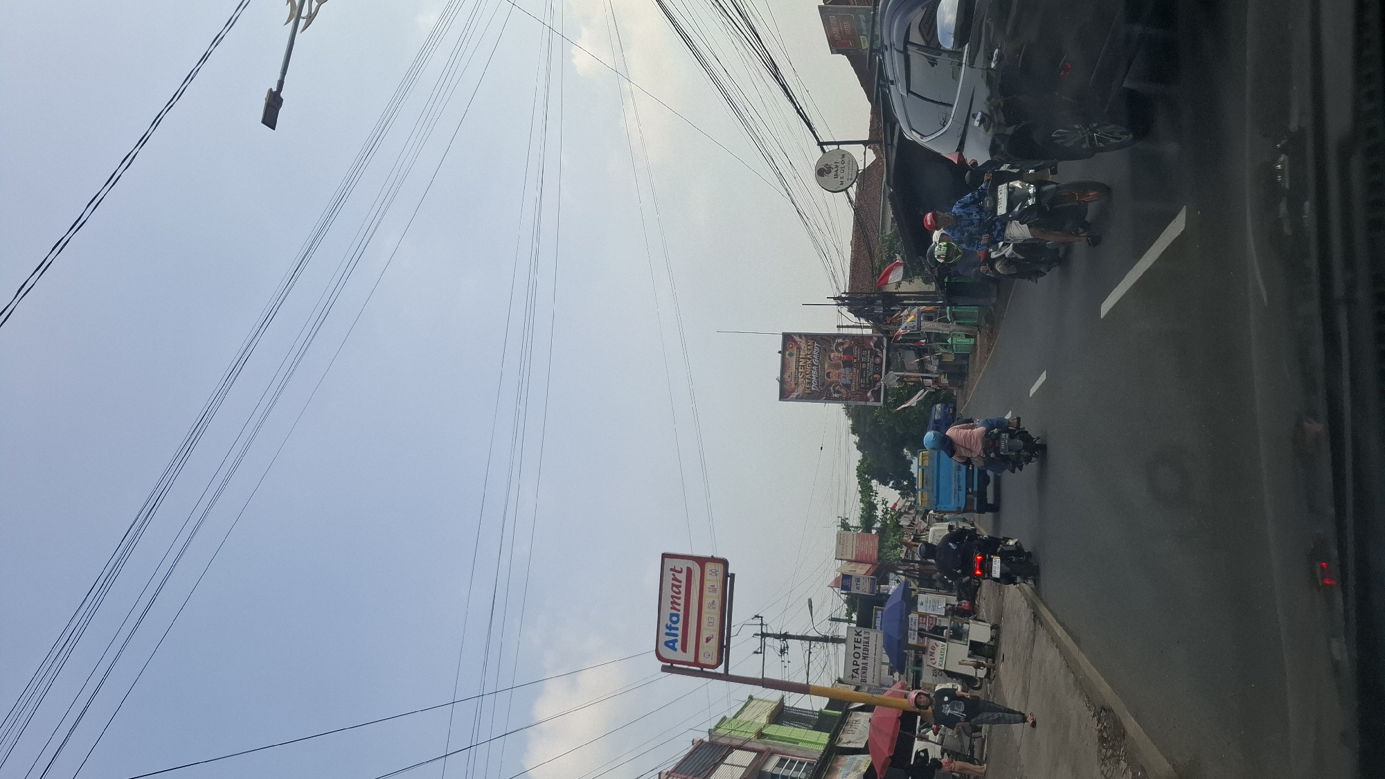 The image captures a street scene in Cilaku, Indonesia, characterized by a moderate flow of traffic under a bright, clear sky. Power lines crisscross the upper portion of the frame, indicative of urban infrastructure. On the left side of the road, a prominent Alfamart sign stands out, suggesting a commercial area. Several businesses, including an apotek (pharmacy), are visible along the sidewalk, with some people walking and standing outside.
The road itself is populated by several motorcycles, some with riders and passengers, navigating the lanes. A blue truck is also present in the traffic. On the right side, a dark-colored SUV is driving alongside the motorcycles. A large billboard advertising an event, possibly a cultural or musical performance, is mounted on a pole further down the street, featuring images of people and text in Indonesian. The overall atmosphere suggests a typical day in a bustling Indonesian town.