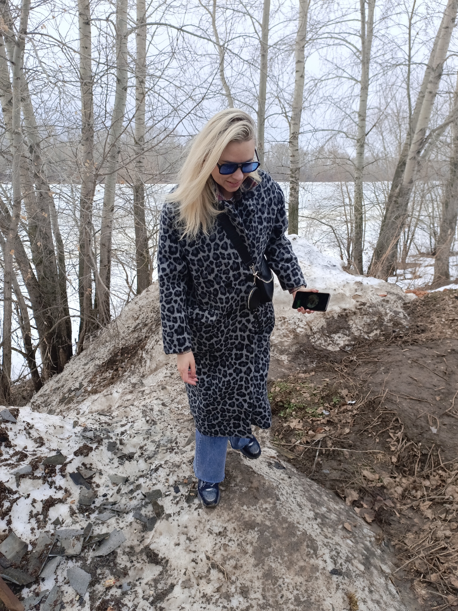 A woman with blonde hair and blue sunglasses stands on a snow-covered dirt mound. She wears a gray and black animal print coat, blue jeans, and dark shoes. She holds a smartphone in her right hand. A black crossbody bag is slung over her shoulder. The background features bare trees, a body of water, and a gray sky. Based on visual characteristics, the location is speculated to be in Chaikovsky, Russia.