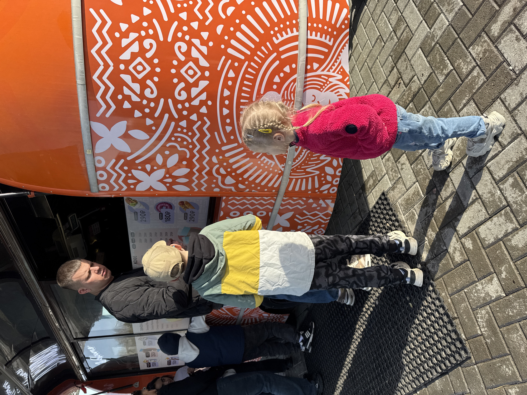 In the Russian city of Perm, a man is observing the menu of a food truck along with two children. The truck is bright orange with a geometric white pattern on its side and lists prices in Russian rubles for items like coffee and donuts. The man is wearing a black jacket and standing close to the service window of the food truck. One child, wearing a jacket with yellow, white, and green panels, stands beside the man. The other child, a girl, is dressed in a pink jacket and blue jeans. The ground is paved with rectangular blocks and a black rubber mat is visible near the truck.