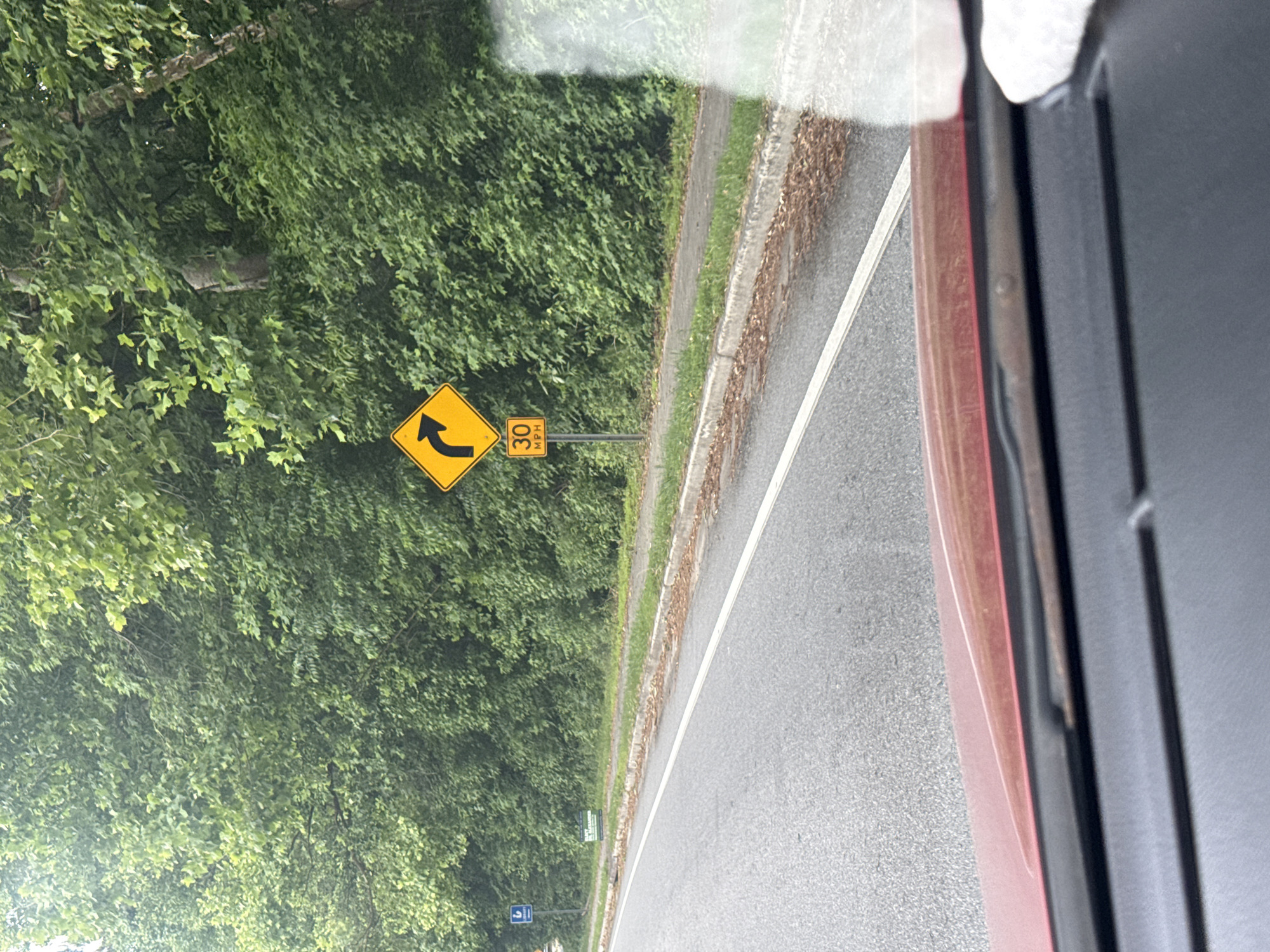 A roadside view from inside a vehicle depicts a curved road sign on the left indicating a left turn ahead, accompanied by a speed advisory of 30 mph. Lush greenery and trees line the side of the road. The location is presumed to be within Mountain Park, United States of America.