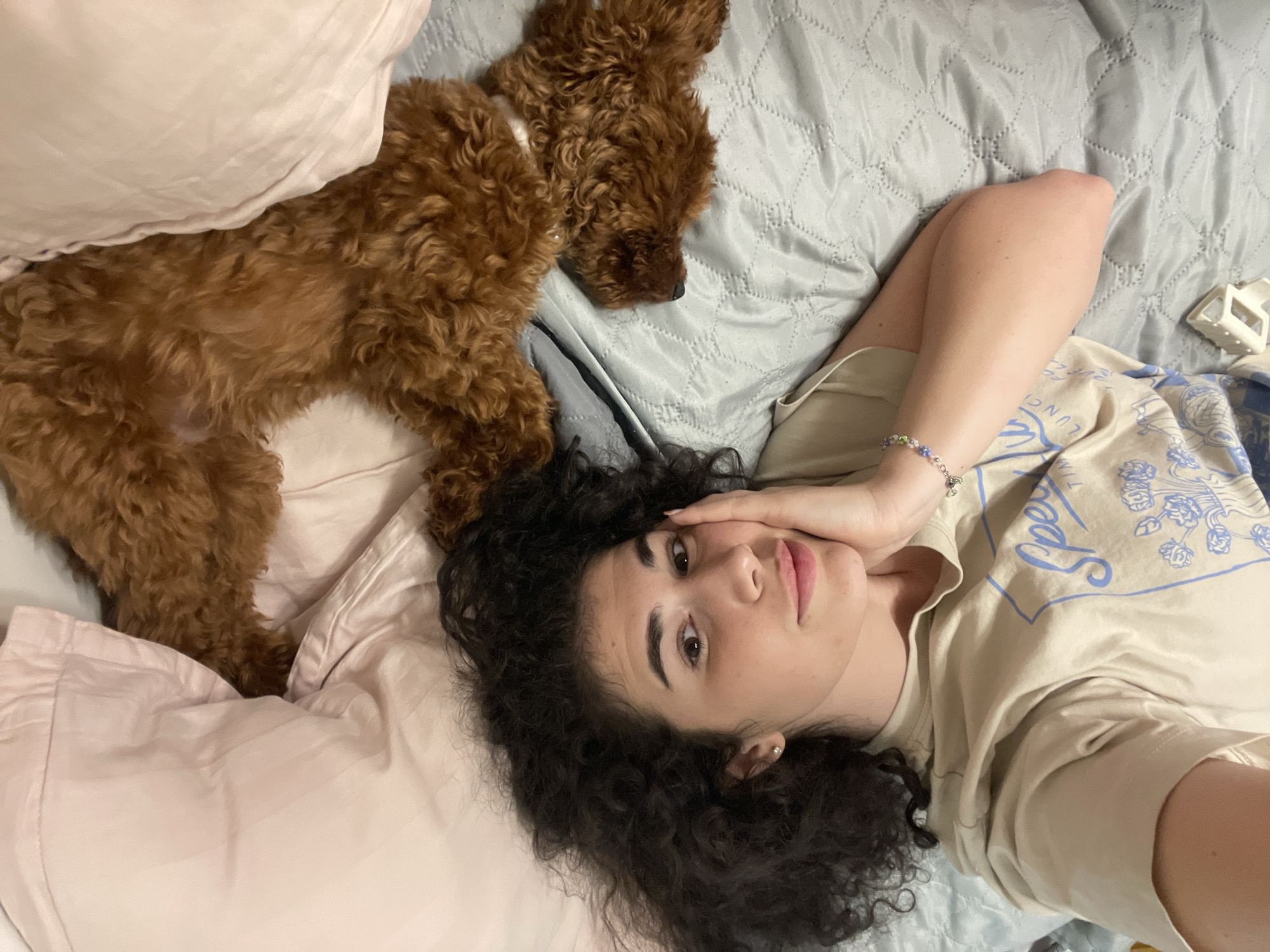 An overhead shot shows a young woman lying on a bed, propped up on her left arm with her right hand on her cheek. She has dark, curly hair and is wearing a beige t-shirt with a blue graphic. To her right, a small, brown, curly-haired dog is also lying on the bed, partially covered by a pink pillow. The bed is covered with a light blue quilted comforter. The scene appears to be indoors, likely in a bedroom. There are no discernible elements in the image that indicate a specific location in Sofia, Bulgaria.