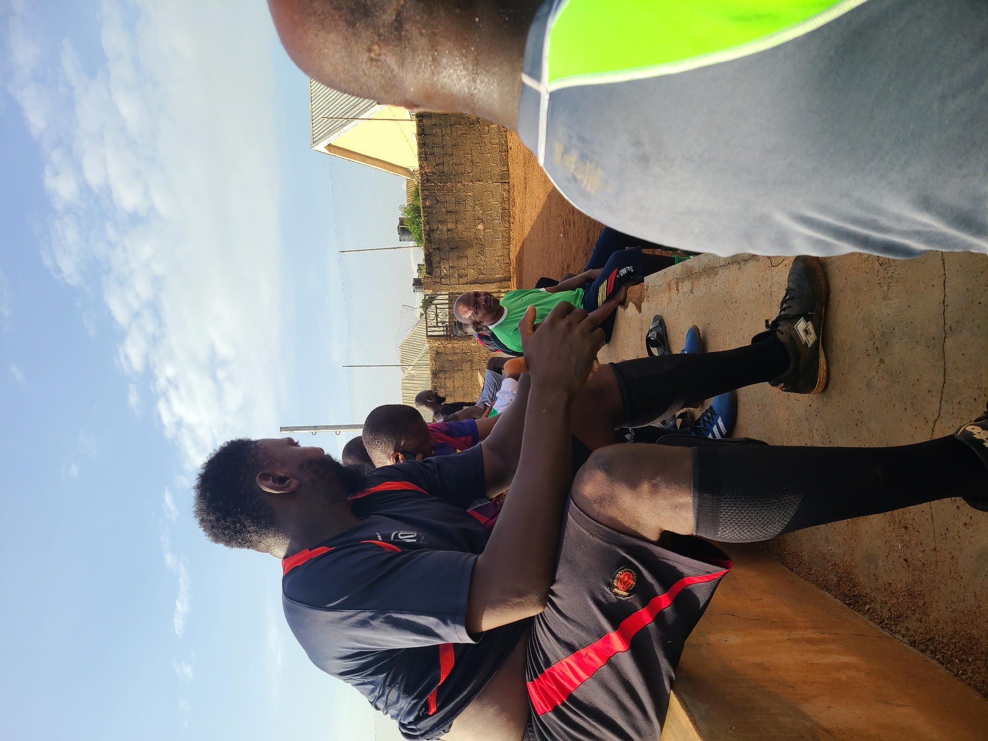 The image shows a group of men sitting on a concrete bench in an outdoor setting, possibly a sports field, in Barakin Lerawi, Nigeria. The man in the foreground is wearing a dark blue shirt with red trim and black shorts with red stripes. He is looking to the left. To his right, other men are seated. One man in the middle ground is wearing a bright green t-shirt and dark pants. The ground is sandy and reddish-brown, typical of the soil in many parts of Nigeria. In the background, there is a brick wall, a chain-link fence, and some buildings with corrugated roofs. The sky is blue with scattered clouds, suggesting it is daytime. The overall atmosphere appears to be casual, possibly after a sporting event or during a break.