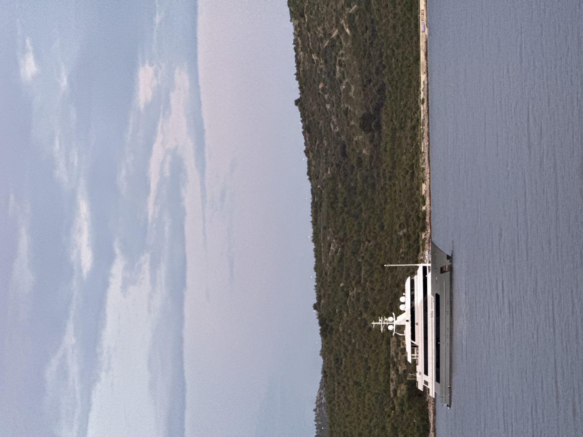 A large, white motor yacht with multiple decks and a sleek design is anchored in the calm, blue waters of the Adriatic Sea. The yacht is positioned horizontally across the frame, with its bow pointing to the right. Behind the yacht, a densely forested, green hillside slopes down towards the water's edge. The sky above is overcast with soft, pale blue and grey clouds. The scene appears to be a serene coastal landscape, possibly near the city of Primošten, Croatia, given its characteristic coastline and vegetation. There are no people visible in the image. The overall impression is one of tranquility and a luxurious maritime setting.