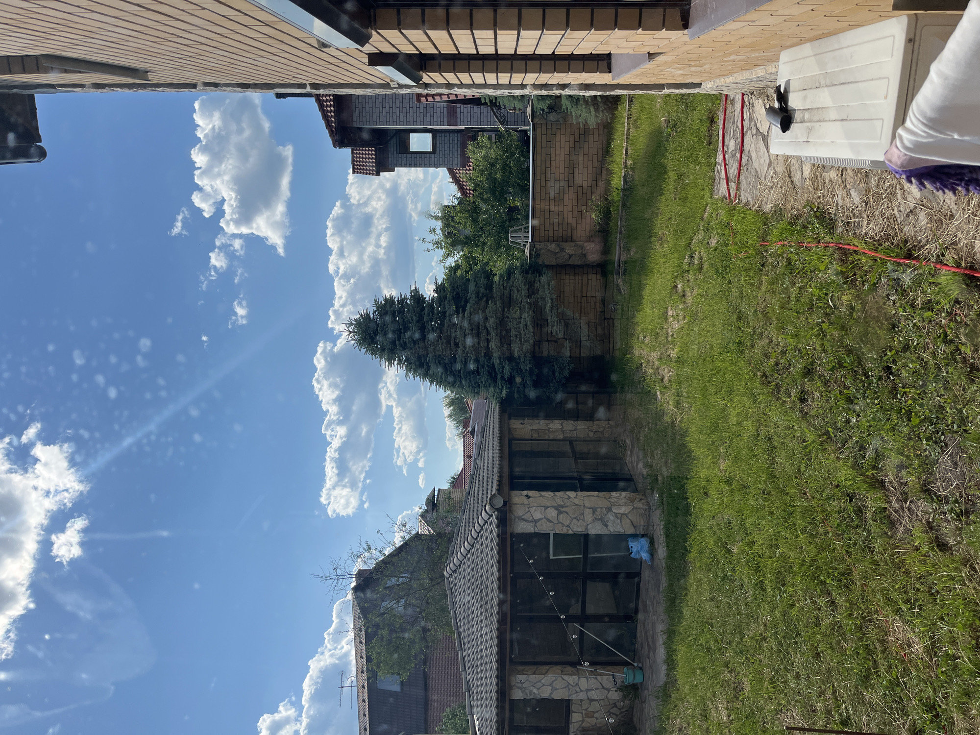 The image depicts a backyard scene on a sunny day in Orekhovka, Russia. In the foreground, to the right, is the side of a light brown brick house with dark framed windows. To the left of the house is a white rectangular object, possibly an air conditioning unit, with a dark object resting on top and a towel draped over its front. A red hose is visible on the ground near the house.
The middle ground of the image features a grassy lawn. In the center of the lawn is a small, stone-clad gazebo with a tiled roof and large glass windows. To the left of the gazebo, a blue plastic bucket and a blue plastic bag are visible. Behind the gazebo, a tall, mature evergreen tree stands prominently. A low, brick wall separates the gazebo area from other parts of the yard.
In the background, several houses with tiled roofs are visible, along with mature trees and foliage. The sky is bright blue with scattered white cumulus clouds. The overall impression is of a residential property on a clear, pleasant day.