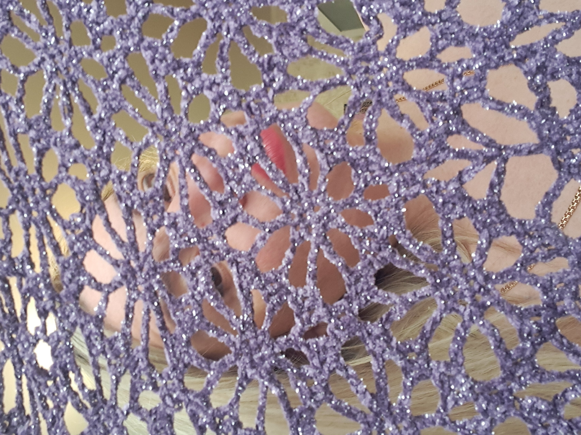 The image shows a person with fair skin and blonde hair partially obscured by a purple, sparkly, crocheted fabric. A hint of red lipstick is visible through the fabric. A gold chain necklace is also visible through the crocheted material on the right side. The background appears to be a light beige or tan color. The image does not provide any information relating to Elektrostal, Russia.