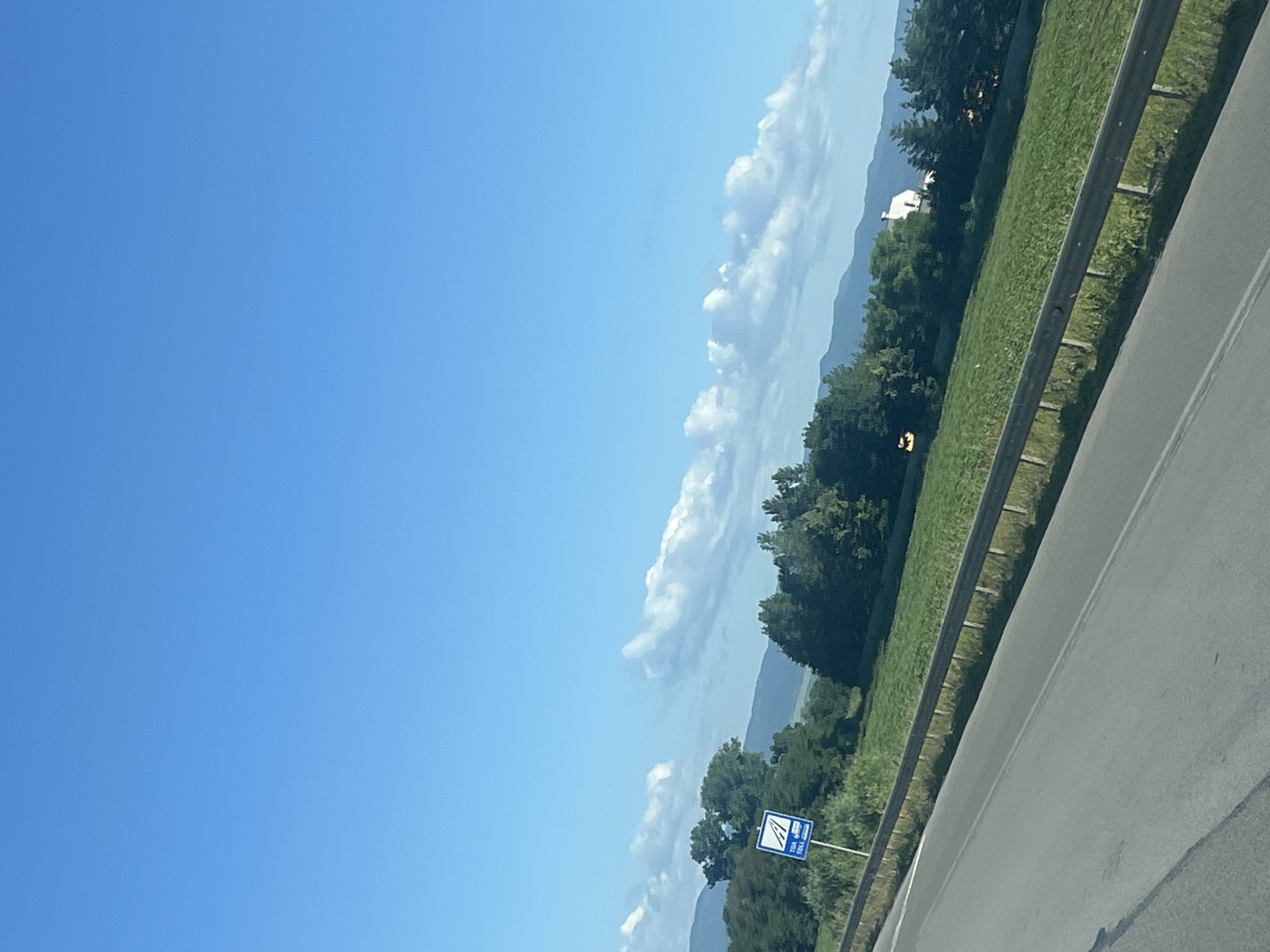 A roadside view captures a clear blue sky with scattered white clouds. In the middle ground, a lush green field is bordered by a dark metal guardrail. A white house is visible nestled amongst a dense line of dark green trees that stretches across the landscape. A blue sign with white text and symbols stands near the left edge of the image, indicating a toll road. The foreground shows a grey asphalt road with white lane markings, suggesting it is a highway. The overall scene depicts a rural landscape under a bright, sunny sky.