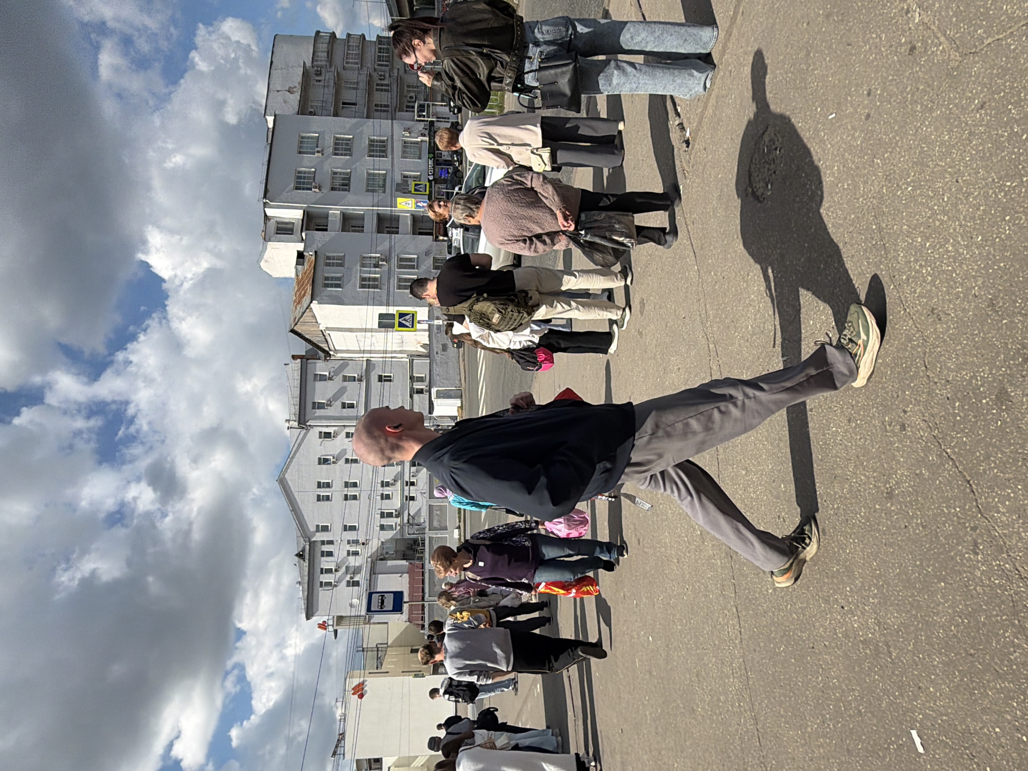 The image depicts a street scene in Nizhny Novgorod, Russia, under a cloudy sky. In the foreground, a bald man in a dark jacket and grey pants is walking towards the right of the frame, casting a prominent shadow on the asphalt. A group of people are visible on the sidewalk and crossing the street in the background. Among them, a woman in a light-colored sweater and dark pants is holding two shopping bags. Another woman in a dark jacket and jeans is taking a picture with her phone. The background shows several multi-story buildings, some with visible windows and balconies. There are also traffic signs and streetlights present. The ground is paved with asphalt, and there are some minor cracks and debris visible. The overall atmosphere appears to be that of a busy urban environment during the daytime.