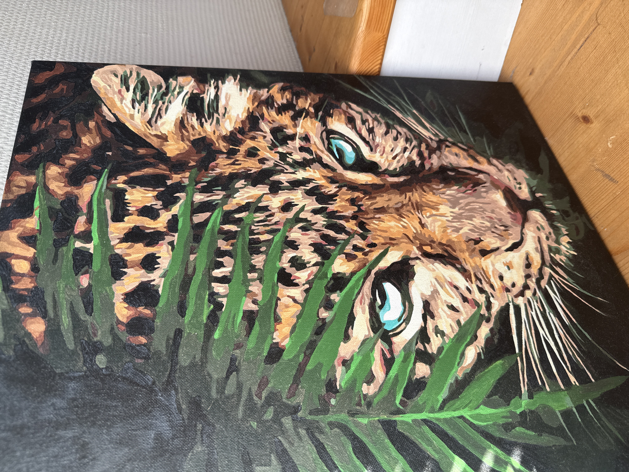 The image shows a close-up view of a painting depicting a leopard partially hidden behind green foliage. The leopard's head is centered, with bright blue eyes gazing forward. The fur is rendered in shades of brown and black with characteristic spots. Green leaves obscure parts of the leopard's face and body. The background is a dark, undefined color. The painting appears to be on a stretched canvas, and the surrounding environment includes a light-colored surface and a wooden structure in the upper right corner. There's no indication of the city of Chaikovsky, Russia.