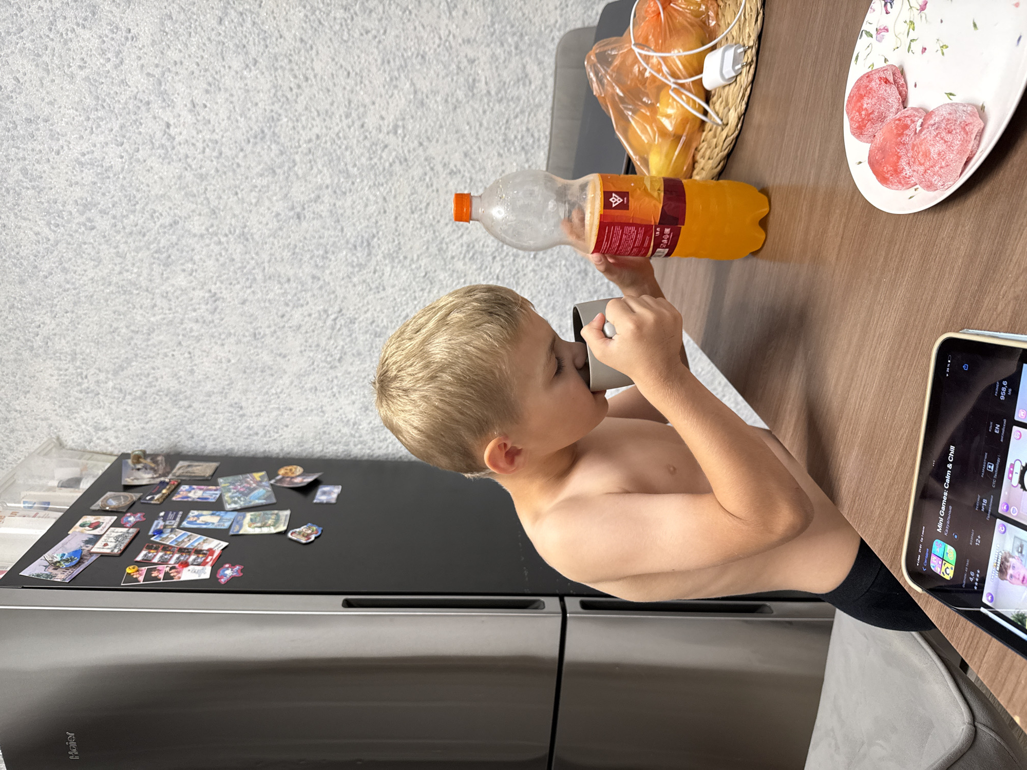 A young boy with blond hair is drinking from a gray mug. He is shirtless and wearing black socks. He is sitting at a wooden table with a bottle of orange soda and a plate of pink, heart-shaped candies. To the left of the boy, there is a black refrigerator with magnets on it. A tablet is visible on the right side of the table, displaying an app called "Mini Games: Calm & Chill". The scene appears to be indoors, likely in a home kitchen. The lighting suggests it is daytime. The boy's expression is neutral as he drinks. The magnets on the refrigerator show various images and designs. The text "Haier" is visible on the refrigerator door. The tablet screen displays "Stary Oskol, Russia" and other app-related information.