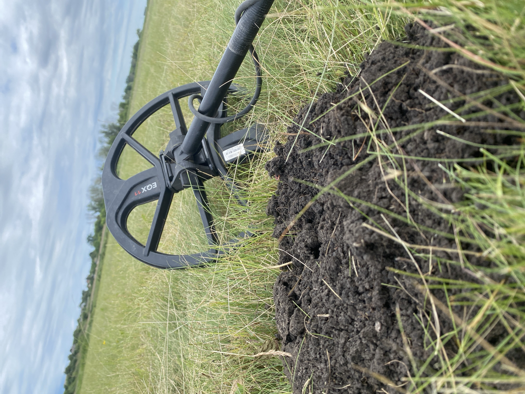 In a grassy field, a metal detector rests on the ground next to a pile of dark soil, suggesting an excavation has taken place. The detector, a black Minelab Equinox 11, is positioned with its coil slightly visible behind the disturbed earth. The surrounding environment is a rural landscape, with a distant treeline and a hazy sky overhead. The location is specified as Ustyvytsia, Ukraine.