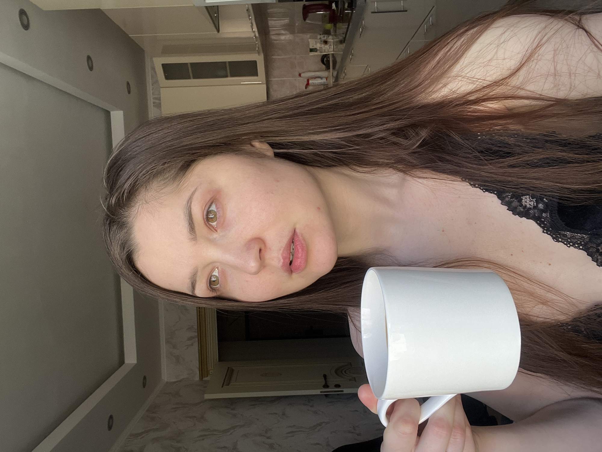 A fair-skinned woman with long, brown hair is holding a white mug in her right hand, likely containing a beverage. She is wearing what appears to be a black lace top. The background shows a domestic interior with a kitchen visible, including cabinets and appliances. A doorway is also visible. The photograph was likely taken indoors, but the precise location in the city of Khimki, Russia cannot be determined without additional information.