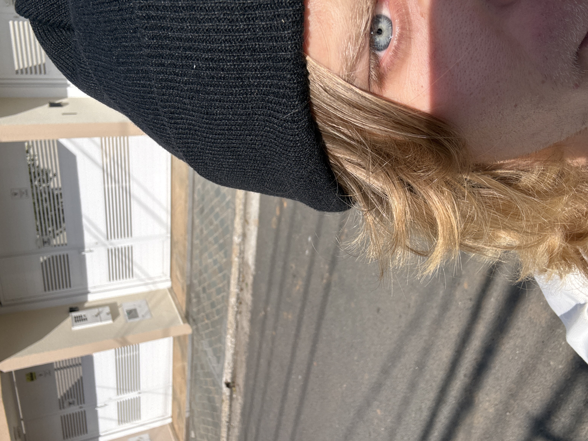This media file is a close-up, Dutch angle photograph of a person's face, focusing on their right eye and blonde hair. The subject is wearing a black beanie that covers their forehead and hair. The person has light skin and blue eyes, with eyelashes and eyebrows visible. A shadow bisects the face, cast by an unseen object, highlighting the texture of the skin. The background features a residential building with white doors and vertical louvers, suggesting a sunlit exterior. Further in the background, a paved street or sidewalk is visible with a brick pattern. The scene is likely outdoors during the daytime, indicated by the strong sunlight and shadows. The overall impression is casual and intimate, a snapshot of a person in an everyday setting. There is no visible text in the image. Given the location context of São Paulo, Brazil, the architecture and lighting are consistent with a location in that region.