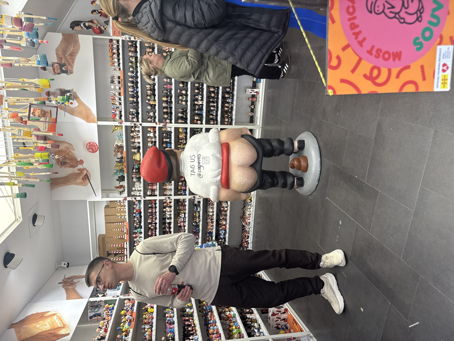 The interior of a souvenir shop, likely located in Barcelona, Spain, specializing in "Caganer" figurines, is depicted. A prominent, oversized statue of a Caganer, a traditional Catalan defecating figure, stands in the foreground, slightly right of center. The statue, facing away from the viewer, wears a red barretina hat, a white shirt with "TAG US @caganers" and an Instagram logo on its back, a red sash, and black pants pulled down to reveal bare buttocks. A sculpted pile of brown feces rests on the grey circular base behind its feet.
To the left of the statue, a young adult male with glasses, a light long-sleeved shirt, dark pants, and white sneakers stands holding a red beverage can, looking towards the left side of the shop. A black watch is visible on his left wrist.
To the right of the statue, two women are partially visible. One woman, with blonde hair tied back, wearing a green jacket, black pants, and Adidas sneakers, stands facing the right, looking at the shelves. Another woman, mostly obscured on the far right, wears a dark puffer jacket and bright blue pants.
The shop walls are lined with numerous white shelves displaying hundreds of smaller Caganer figurines, organized into categories such as "ART I CULTURA" and "POLITICS." Above the shelves, large wall art depicts hands engaged in crafting or painting figures. Colorful paintbrushes hang from the ceiling. The floor is covered in dark grey tiles. A yellow and orange sign, partially visible in the bottom right, reads "MOST TYPICAL SOUVE..." and identifies itself as an "OFFICIAL TOURISM PRODUCT."