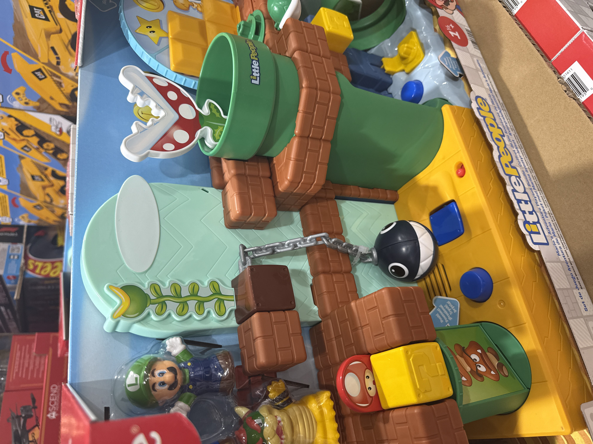 The image shows a "Little People Mario's Adventure Playset Bundle" toy set still in its packaging. The set features various elements from the Super Mario video game franchise, including a green pipe with a Piranha Plant, a figure of Luigi, a Chain Chomp, and other building blocks and characters. The packaging is lying on a surface that appears to be cardboard, possibly in a retail setting such as a warehouse or store. The text on the box is partially in English, French, and Spanish. There are other retail products visible in the blurred background. There is no reference to Puebla, Mexico.
