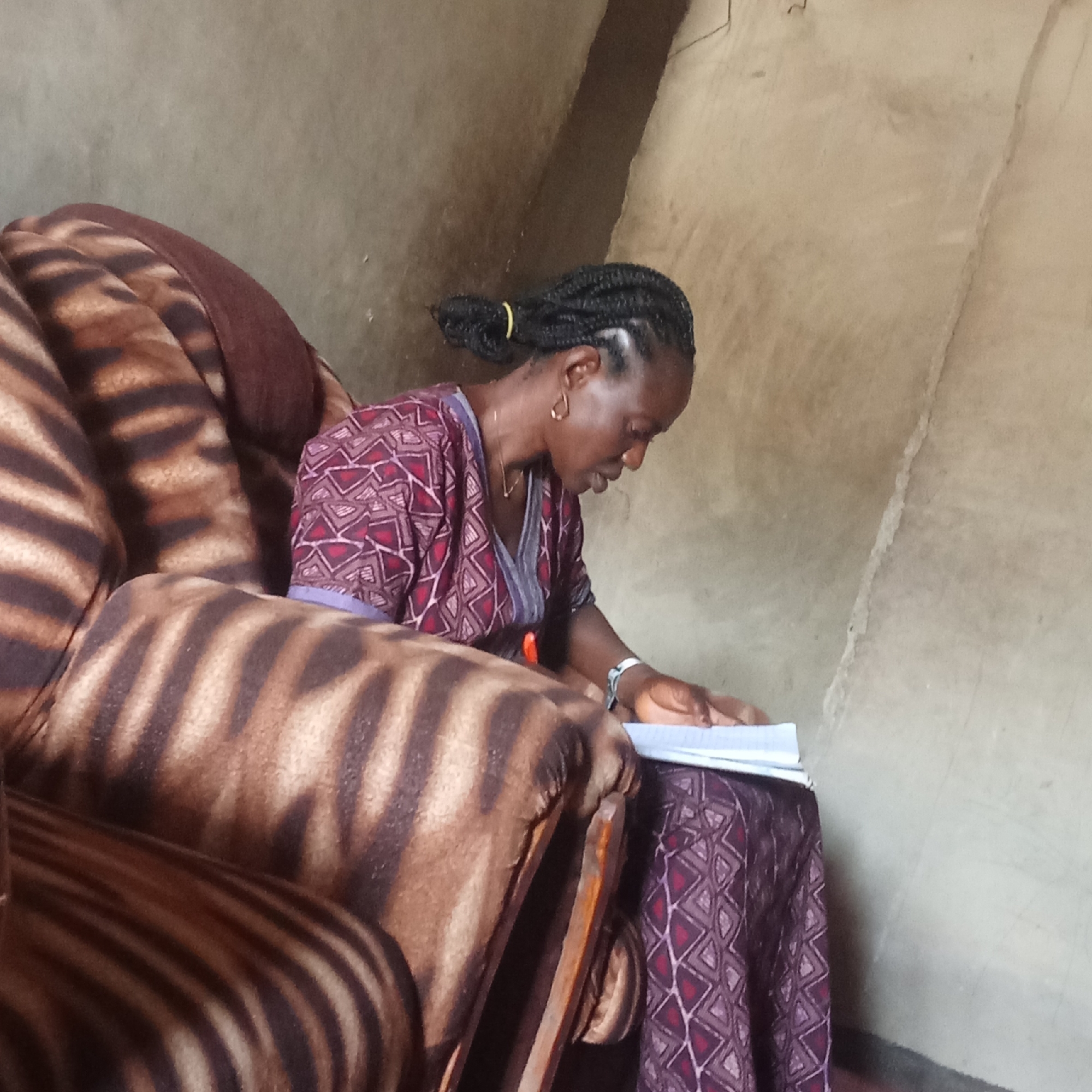 In Akwanga, Nigeria, an adult Black woman sits in an oversized armchair with a zebra-striped pattern. The chair is against a light beige wall with minor imperfections. The woman has braided black hair pulled back with a yellow hair tie, and is wearing a patterned purple and burgundy dress. She appears to be reading or writing in a notebook, looking down at the pages with a focused expression. She is wearing earrings and a watch. The room is simply furnished.