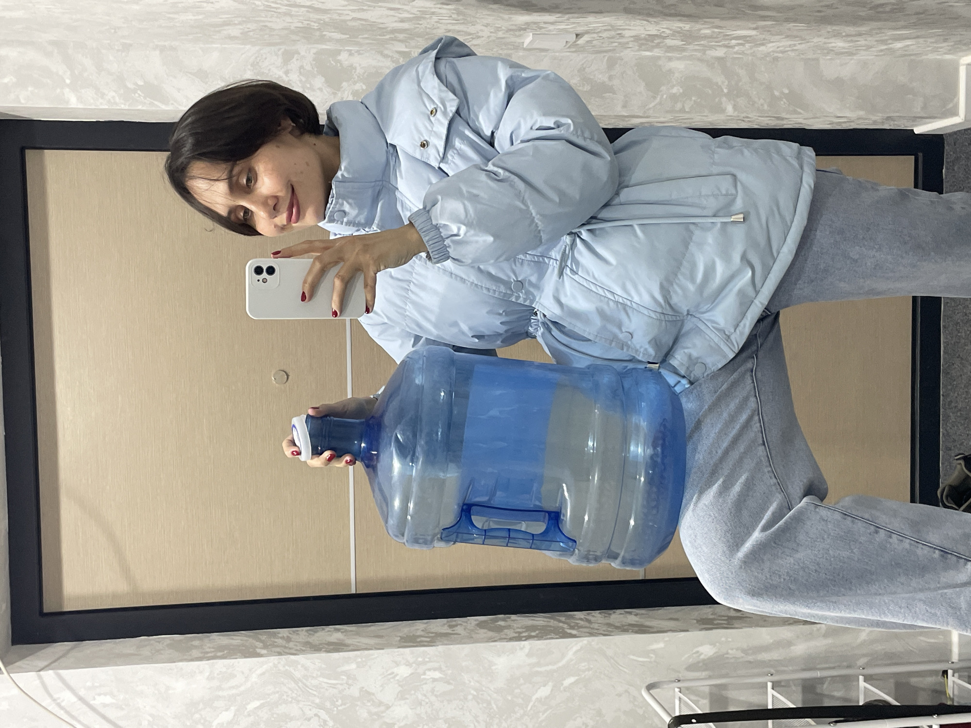 A young woman with short, dark hair is indoors, holding a large blue water jug in one hand and a white smartphone in the other. She is taking a selfie. She wears a light blue puffer jacket and light gray jeans. The background shows a wall with a dark-framed beige panel, suggesting an office or similar interior space. The presence of what appears to be a clothes drying rack in the bottom right corner suggests the space may have dual functionality. There is insufficient information in the image to determine if the location is in Izhevsk, Russia.
