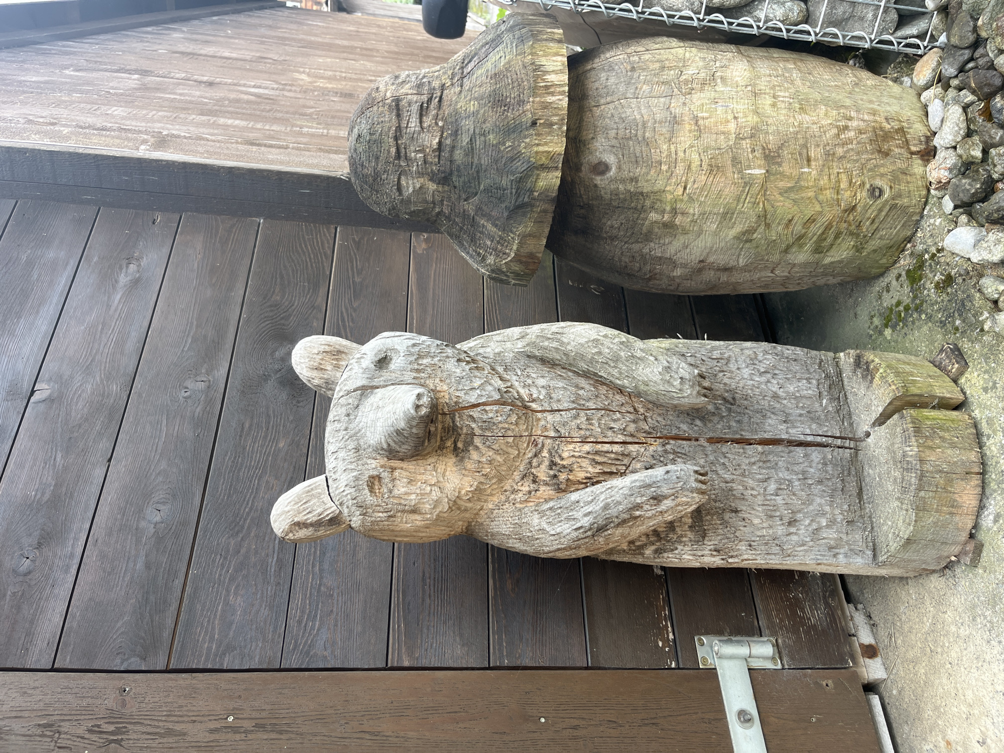A wooden sculpture of a bear stands to the left of a wooden structure. The bear sculpture is carved from a single piece of wood and stands upright on a wooden base. It has carved ears, eyes, and a snout. To the right of the bear is another wooden sculpture that resembles a mushroom. This sculpture is also carved from wood and is positioned on a circular wooden base. The wooden structure behind the sculptures appears to be a building, with horizontal wooden planks. In the foreground, there is concrete and gravel. The overall scene suggests an outdoor decorative display, possibly in a garden or a public space. The location is Wengen, Switzerland.