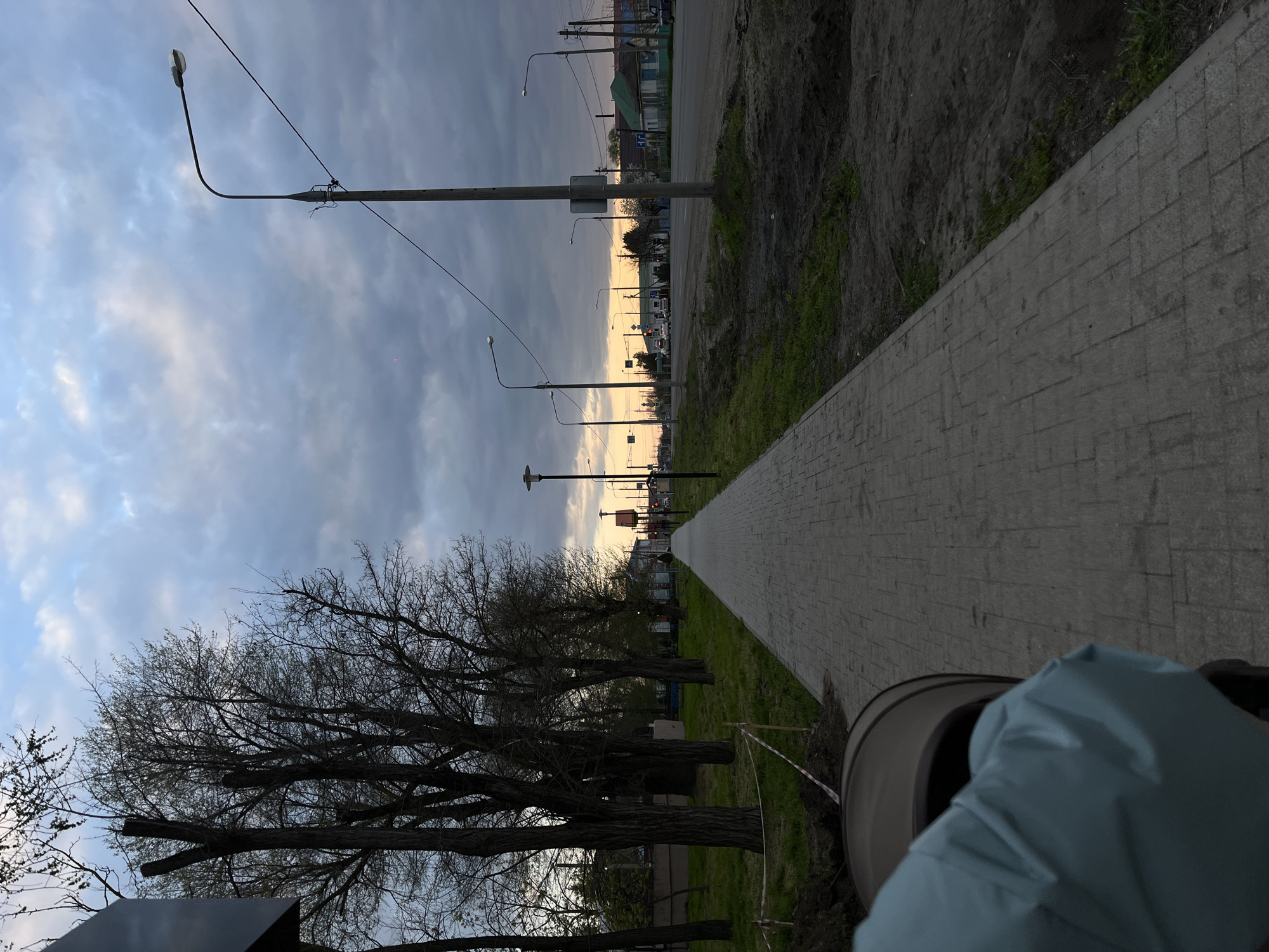 The image shows a paved walkway in Novopokrovskaya, Russia. The walkway extends into the distance, flanked by grass and bare trees on one side and a dirt patch and street on the other. Streetlights line the walkway. The sky is cloudy with a hint of sunset. A piece of equipment is partially visible in the foreground.