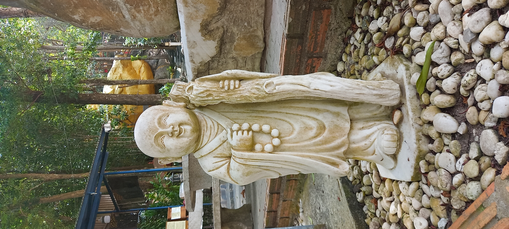A white stone or marble statue of a young Buddhist monk, possibly a novice, stands upright in an outdoor setting in Moo 7, Thailand. The monk statue has a smooth, slightly weathered texture and displays a serene, smiling expression on its face. It holds a textured, bark-like staff or tree trunk with its right arm, and a string of prayer beads in its left hand. The statue wears simple robes with flowing folds.
The statue is positioned on a rectangular base, which is surrounded by numerous light-colored, rounded pebbles covering the ground. In the background, lush green trees and foliage are visible under bright daylight, suggesting a sunny day. Further back, hints of golden-yellow structures, possibly other decorative elements or parts of a building, can be seen amidst the greenery. A dark metal fence or barrier is also present in the middle ground, along with some reddish-brown bricks or tiles on the ground or as part of a structure. The overall scene conveys a peaceful and spiritual garden or temple atmosphere. No text is visible in the image.