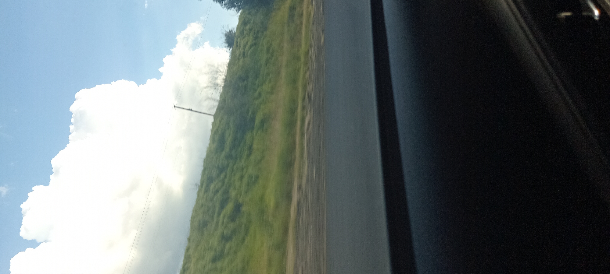 The image is a vertical shot taken from inside a vehicle looking out towards a cloudy sky and a grassy hillside. In the foreground, the dark interior of the car is visible, likely the dashboard or door panel. A grey road is visible in the middle ground, with a blur indicating motion. The background shows a green, grassy hillside with a few sparse trees and what appears to be a telephone pole. The sky is predominantly blue with a large, bright white cloud on the left side of the frame. The photo was taken in Valky, Ukraine.