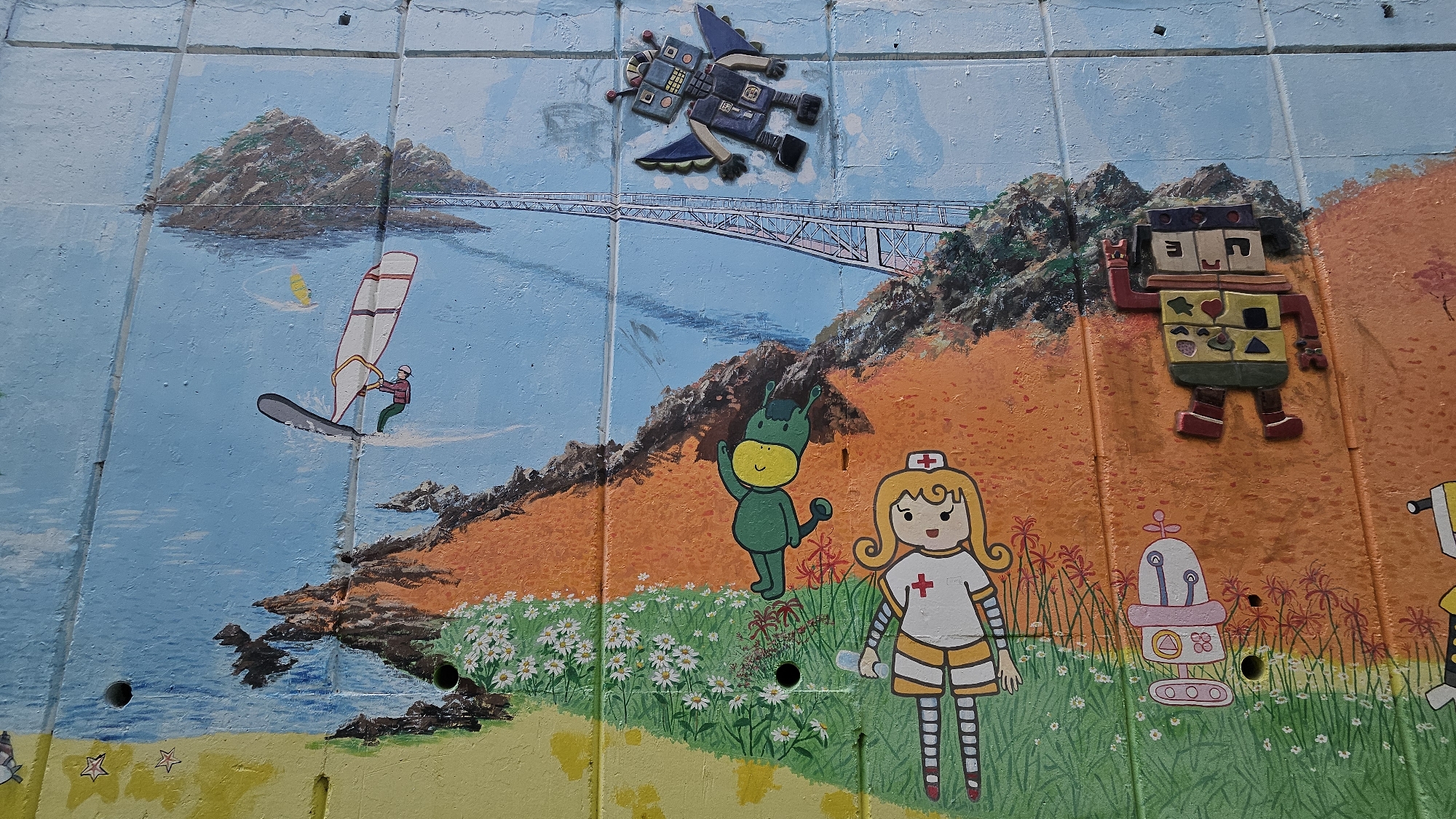 This is a close-up, slightly high-angle photograph of a painted mural on a concrete wall. The mural depicts a whimsical seaside scene with several cartoonish figures.
In the top left, there is a painting of a body of water with a person windsurfing. A small island with a bridge is visible in the background. Above the bridge, a three-dimensional, mosaic-style flying robot is affixed to the wall.
To the right, the scene transitions to an orange landmass with green grass and white daisies in the foreground. Cartoon characters, including a green monster, a nurse with a red cross on her uniform, and various robots are painted and affixed to the wall.
The mural is painted on concrete blocks, the seams of which are visible. There are also several small holes in the concrete wall, disrupting the painted scene.
Based on the mural's content and known facts, this photograph was taken in Ulsan, South Korea.