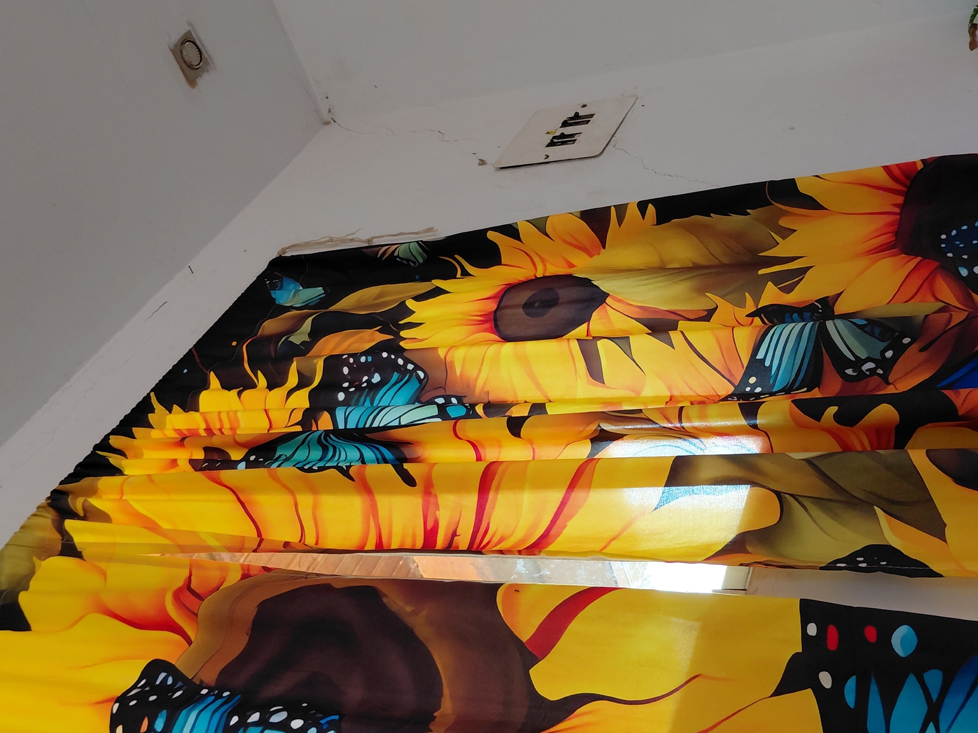 The image shows a close-up view of a vibrant yellow curtain with a sunflower and butterfly pattern. The curtain is hanging in what appears to be a room with white walls. A light fixture is visible on the ceiling, and a light switch or outlet cover is mounted on the wall. The overall impression is of a domestic interior. The image is a digital photograph taken with a mobile device.