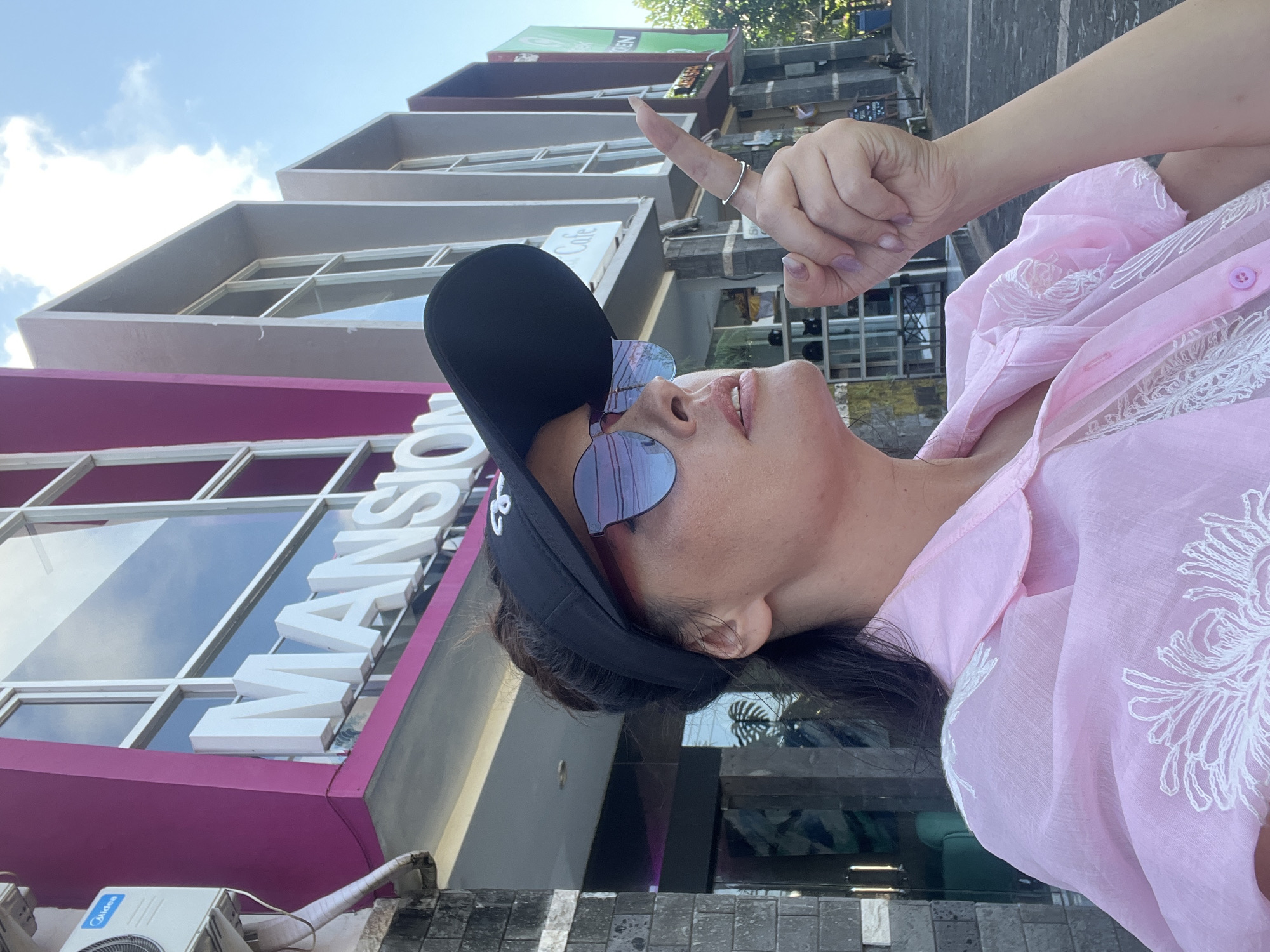 A woman wearing sunglasses, a visor, and a pink shirt with white embroidery is pointing up and to the right in front of a building with "MANSION" in large white letters across its facade. The building is located in Kuta Selatan, Indonesia. The sky is clear and blue with some white clouds. The building has a modern architectural style with large windows and a pink accent color.