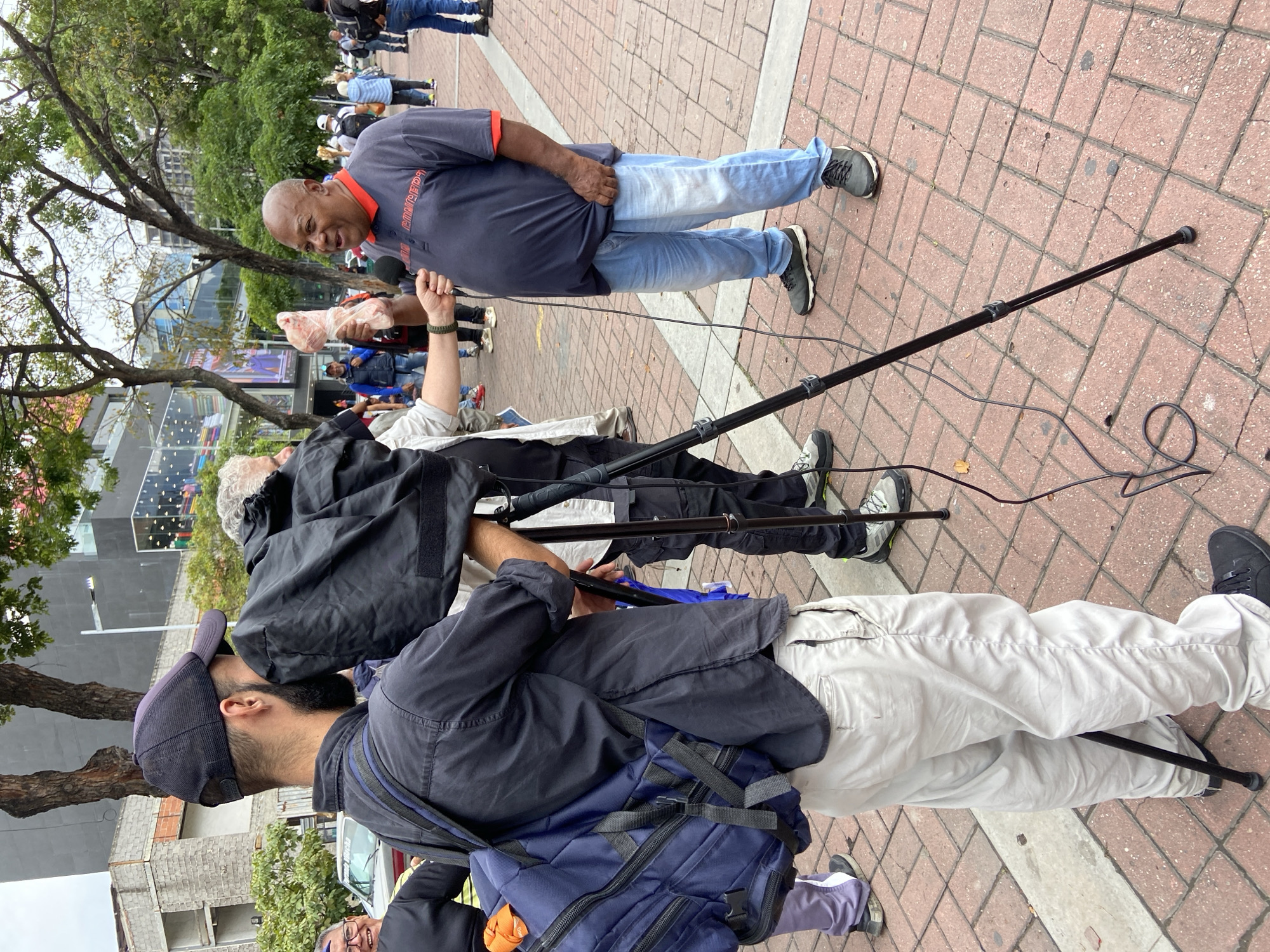 This outdoor image, taken in Caracas, Venezuela, during the daytime under what appears to be an overcast sky, captures a public address or media event. The central figure is a bald, dark-skinned man wearing a dark blue polo shirt with an orange collar and light blue jeans. He is actively speaking into a microphone held in his left hand. Strikingly, he holds a piece of raw meat, possibly chicken, wrapped in clear plastic, in his right hand.
He is surrounded by individuals who appear to be part of a media crew. One person, with white hair and wearing a black jacket, is operating a long black boom pole, directing it towards the speaker to capture audio. Another crew member, visible in the foreground with a dark cap and black shirt, is also holding the boom pole and carrying a dark blue backpack. Black cables from the audio equipment trail across the red brick pavement.
In the background, a crowd of people is visible, indicating a public gathering. The setting includes mature trees and urban buildings, some appearing under construction. The ground is paved with reddish-brown bricks. On the speaker's shirt, the text "EL VERDADERO" is partially visible. The scene conveys an engaged speaker delivering a message, possibly using the raw meat as a symbolic prop.