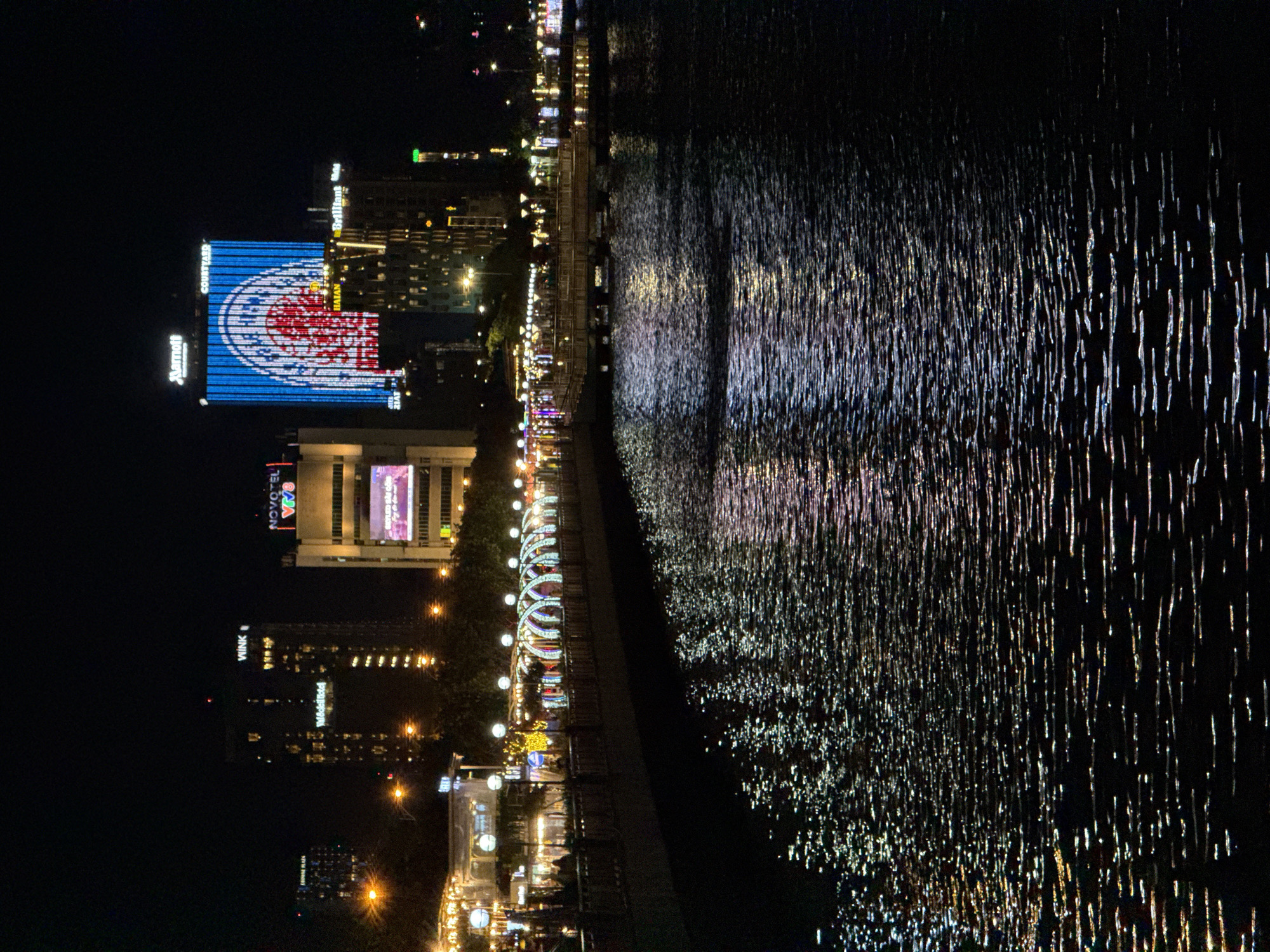 The image depicts a nighttime scene along a waterfront in Da Nang, Vietnam. The foreground is dominated by the dark water of a river or bay, reflecting the vibrant lights of the city. The reflections create a dazzling, shimmering effect on the water's surface, appearing as streaks of white and colorful light.
Along the waterfront, a promenade is visible, lined with buildings and illuminated by decorative lights. Many of these lights are strung along arches, creating a festive atmosphere. Several buildings are lit up, with some featuring large LED screens displaying advertisements or information. One prominent building has a large, blue and red illuminated graphic on its facade, with text that appears to read "COURTYARD" and "Marriott" on the left side of the graphic, and "VIVA" and "NOVOTEL" on another building further down the waterfront. Another building shows "WINK" and "Woodland". The overall impression is one of a lively urban environment at night. The time of day is clearly night, and there are no indications of specific weather conditions. The scene conveys a sense of urban vibrancy and nighttime activity.