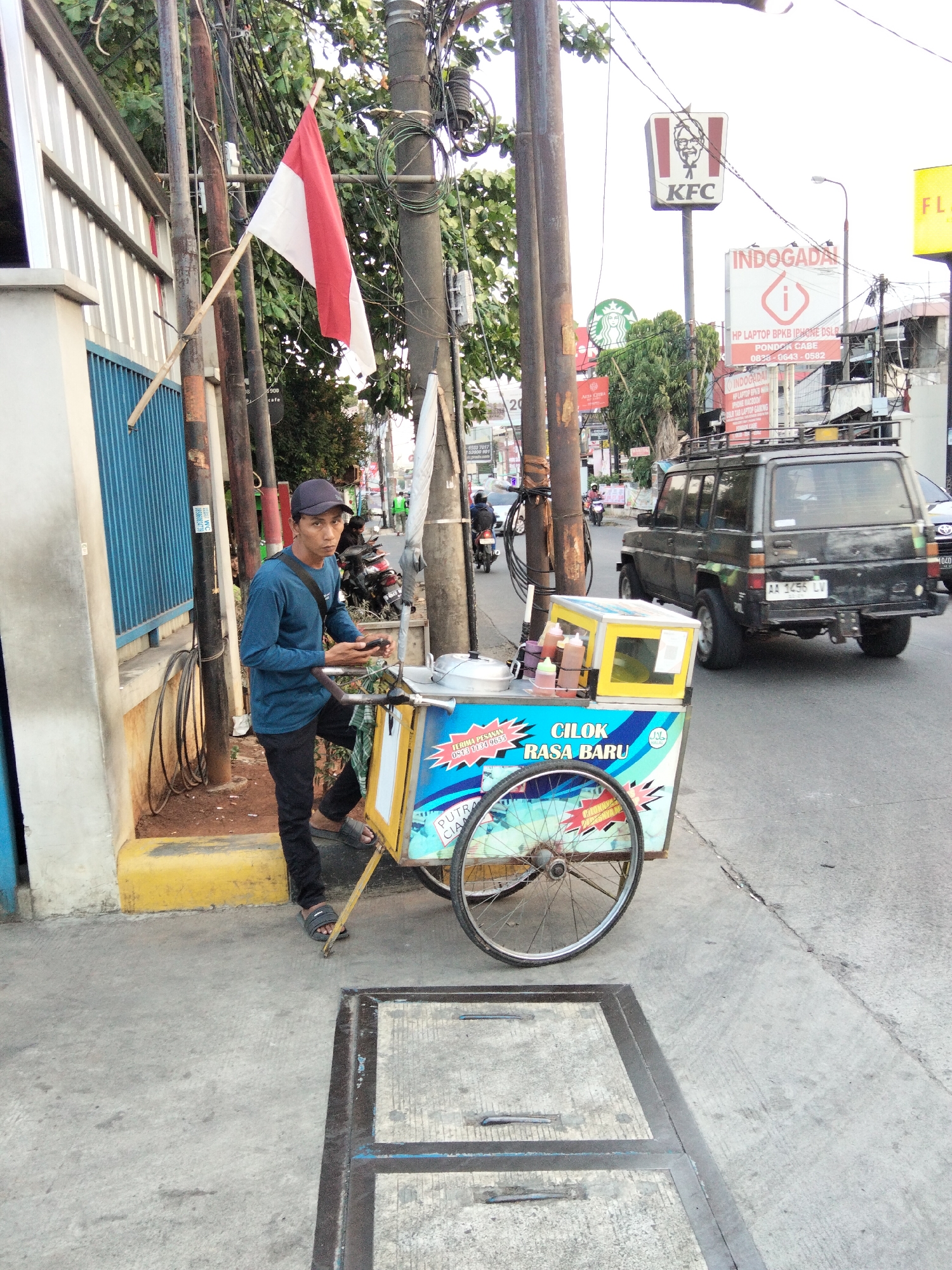 A street vendor stands next to a food cart on a sidewalk in South Tangerang, Indonesia. The vendor, a male wearing a blue long-sleeved shirt, dark pants, a dark cap, and sandals, is looking towards the camera while holding a mobile phone. The food cart is yellow and blue with text that reads "CILOK RASA BARU" and "TERIMA PESAN". It has bottles of sauce on top and a large wheel. Behind the vendor, a red and white Indonesian flag is mounted on a pole. In the background, there are several utility poles with wires, trees, a KFC sign, an INDOGADA sign, and a black SUV driving on the road. The ground is paved with concrete, and there is a metal grate in the foreground. The environment appears to be an urban street scene.