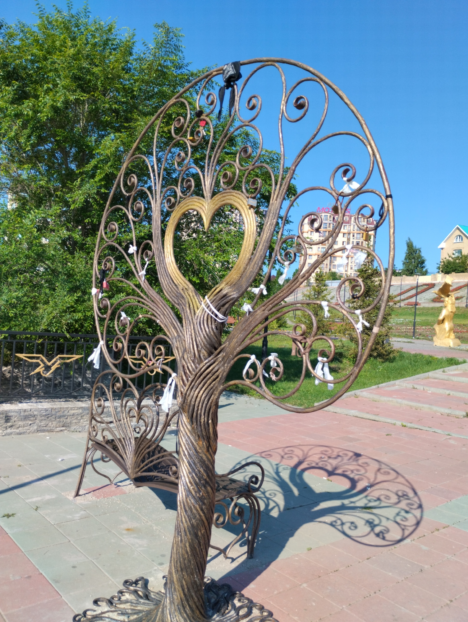 The image shows a decorative metal tree sculpture with a heart shape at its center, located in a city park in Kostanay, Kazakhstan. The sculpture is made of twisted metal and has small white ribbons tied to its branches. The tree is positioned on a paved pathway with brick and concrete tiles, and its shadow is cast on the ground. In the background, there is a black metal fence with gold decorative elements, lush green trees, and modern multi-story buildings. To the right of the tree sculpture, there is a golden statue of a person or eagle on a pedestal and a green-roofed building. The sky is clear and blue, indicating a sunny day. The overall scene depicts an outdoor public space, possibly a square or a promenade, adorned with artistic installations.