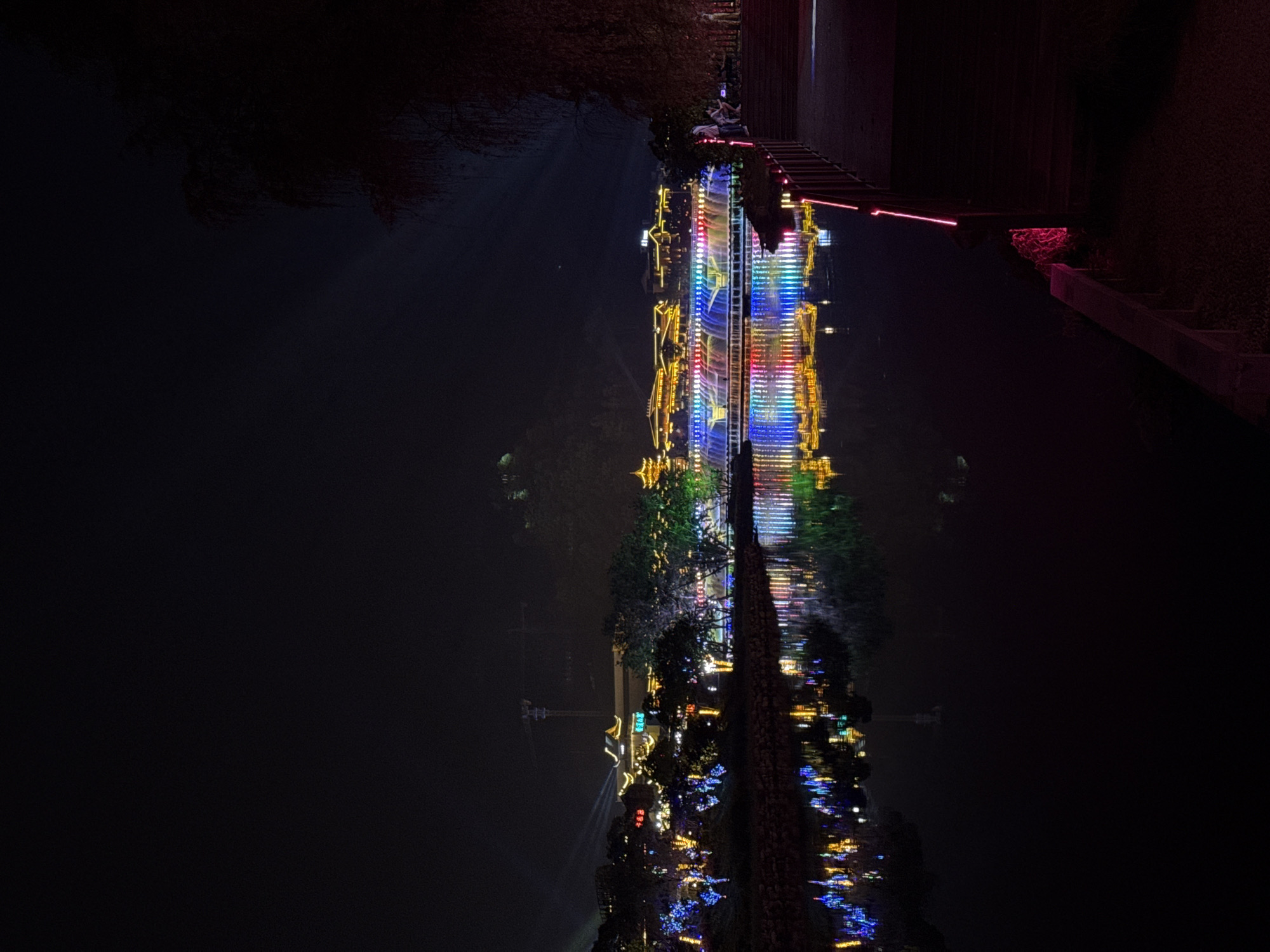 The image captures a night scene along a waterway in Wulingyuan, China. Reflections of illuminated buildings and trees are visible in the dark water, creating a mirror-like effect. The buildings display colorful lights, including blue, red, and yellow hues. Trees line the waterfront, adding to the overall aesthetic. In the upper right corner, a staircase and a portion of a wall are visible, subtly lit with red lighting. The opposite bank is shrouded in darkness, with only faint outlines of vegetation discernible. A crane is noticeable in the distance. The overall atmosphere is serene and urban at night.