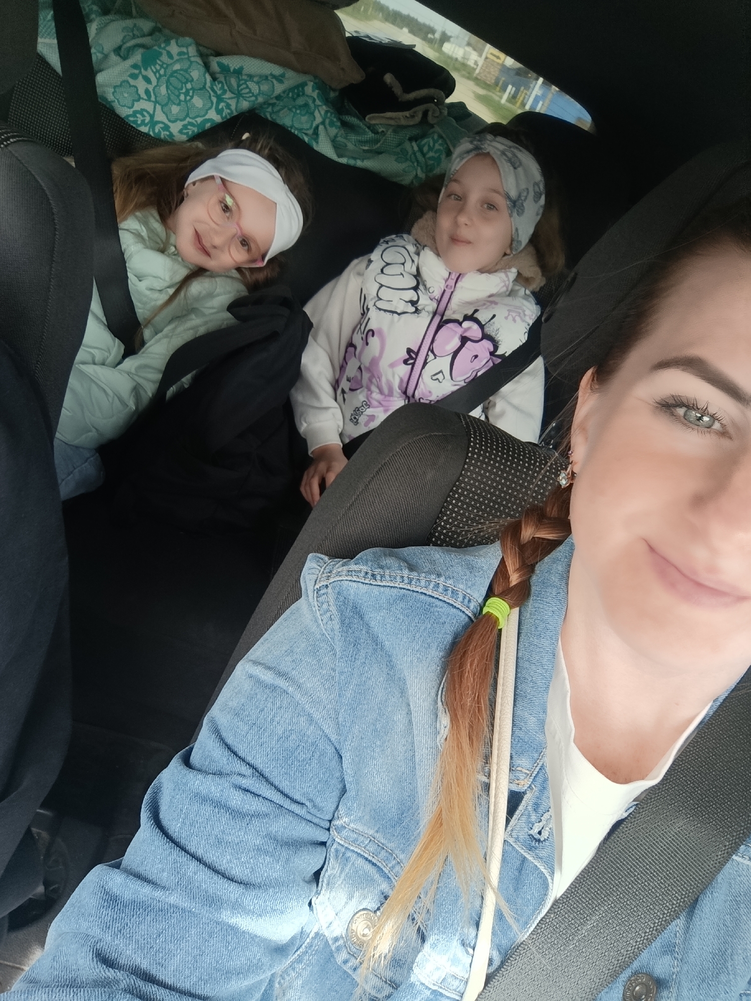 The interior of a vehicle is shown in this medium shot taken in Unknown, Unknown. In the back seat are two young girls wearing headbands and jackets, both secured with seatbelts. The girl on the left wears glasses with pink frames. They are surrounded by blankets and bags. In the front seat, a woman wearing a denim jacket is also wearing a seatbelt. She is smiling and looking at the camera. Her red-brown hair is in a braid with a green hair tie. The background shows a blurred view of what is outside.