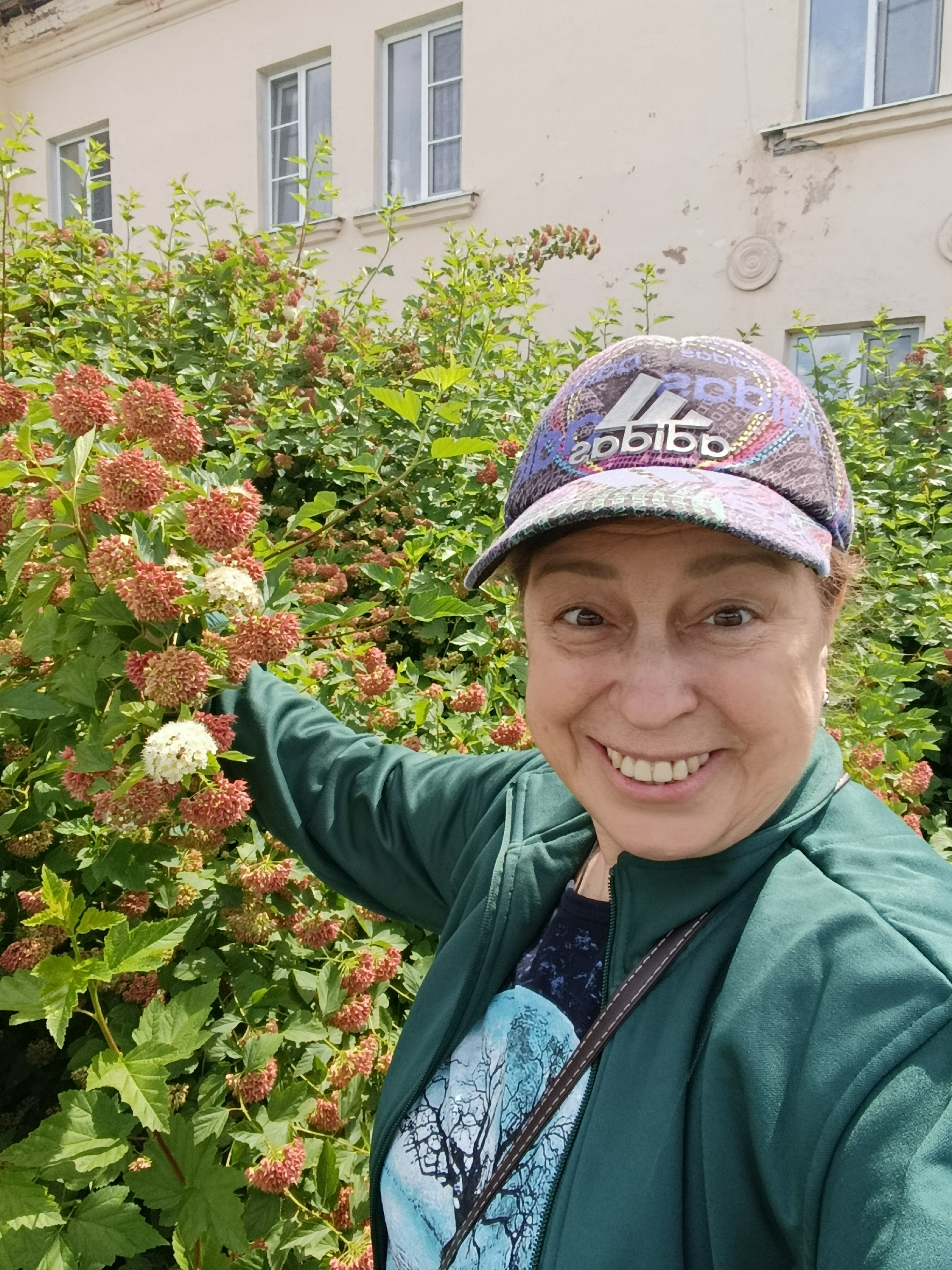 In an outdoor setting, a woman is captured in a selfie, standing in front of a large bush with clusters of pink and white flowers. She is wearing a green jacket, a dark t-shirt with a nature-themed graphic, and a colorful baseball cap with the Adidas logo. The woman is smiling broadly, her right hand is extended towards the bush, pointing at a cluster of flowers. In the background, a light-colored building with several windows and some architectural details is visible. The overall scene suggests a casual, pleasant day, possibly in a park or garden.