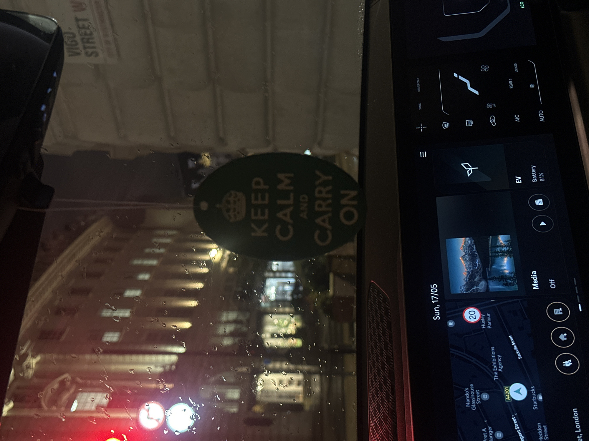 The image shows a view from inside a vehicle, possibly a car, on a rainy day. Through the water-covered windshield, a building with multiple windows is visible. A "Keep Calm and Carry On" sticker is attached to the windshield. To the right of the windshield is a digital display, showing elements like a map indicating the location in London, United Kingdom, near Regent Street, Heddon Street, and Sackville Street. The map also indicates the presence of a Starbucks. The display also indicates the date as Sunday, May 17th, shows the car's battery level at 81%, and includes climate control settings. A sticker on the windshield reads "VIGO STREET W".