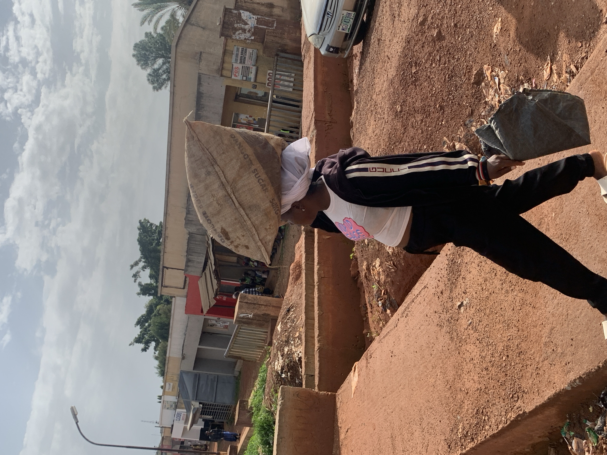 The image depicts a street scene in Gura Top, Nigeria. A person is walking from left to right, carrying a large sack of goods balanced on their head. The sack appears to be made of burlap and has some visible text, possibly indicating the contents are sugar. The individual is wearing a white t-shirt with a pink design, black pants, and a black jacket with white and red stripes on the sleeves. A white cloth is wrapped around their head, under the sack. In their right hand, they are holding a folded grey bag. The environment is characterized by a dirt road with a raised concrete walkway on the left. In the background, there are several buildings that appear to be shops or market stalls, some with corrugated metal roofs. A street lamp is visible on the left. A silver car with a Nigerian license plate is parked on the right side of the road. The sky is partly cloudy. The ground is covered in red earth and some debris. Several other people are visible in the background near the shops.