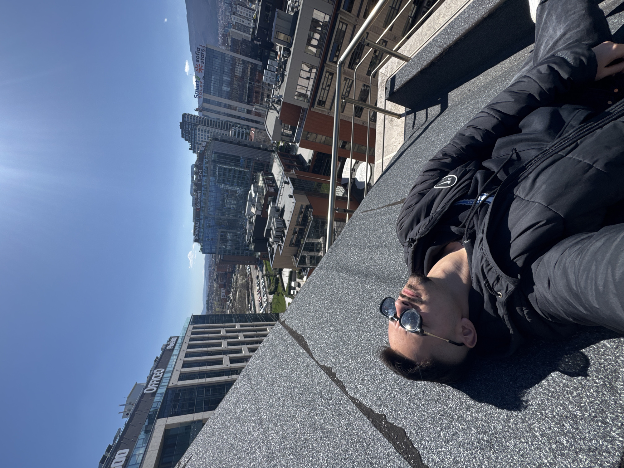 A man wearing sunglasses and a dark jacket is lying on a flat rooftop with a view of Sofia, Bulgaria. The buildings visible in the background include Office 1 Superstore and the Sofia Stars Business Center. The sky is blue.