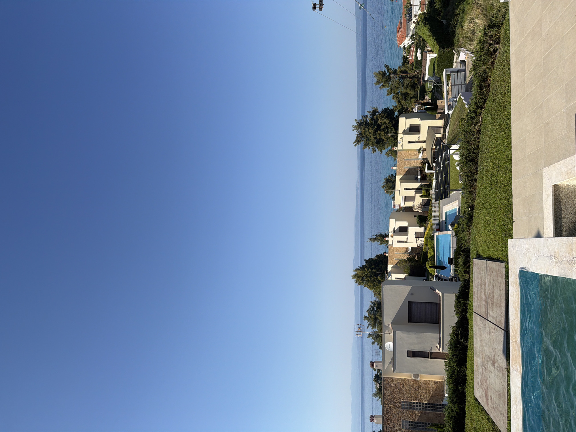 This is a vertical, full shot of a modern resort complex located in Pallini Municipal Unit, Greece. The scene captures several multi-story buildings with white facades and flat roofs, nestled amongst lush green landscaping. There are at least two swimming pools visible, one with bright blue water and lounge chairs nearby, and another with darker, rippling water in the foreground. The buildings appear to be set on a slope, leading down towards a body of water, likely the sea, which stretches to the horizon under a clear, bright blue sky. Trees and manicured hedges dot the landscape, creating a serene and upscale atmosphere. A cable car system is visible in the upper portion of the frame, suggesting the resort might be located near a mountain or a scenic viewpoint. The time of day appears to be daytime, with ample sunlight illuminating the scene. There is no visible text.