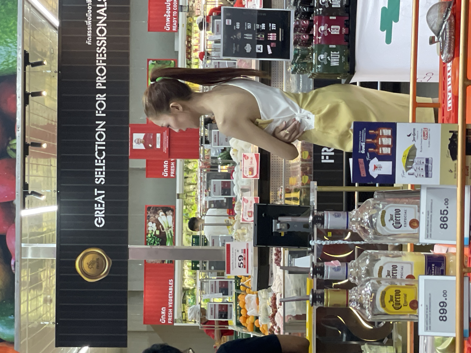 The image captures a young woman with her hair in a ponytail, wearing a white top with a single strap and a yellow skirt, standing in a supermarket aisle in Pattaya City, Thailand. She is facing towards the right side of the frame, with her arms crossed. Behind her, shelves are stocked with various products, including vegetables, fruits, and bottles of alcohol. In the foreground, there are three bottles of Jose Cuervo Especial tequila displayed, with price tags below them. One bottle is yellow, and the other two are clear. To the right, there are shelves with bottles of various liquors, including brands like White, Red, and Dark. A large banner above the shelves reads "GREAT SELECTION FOR PROFESSIONALS". The environment appears to be a well-lit, modern supermarket.