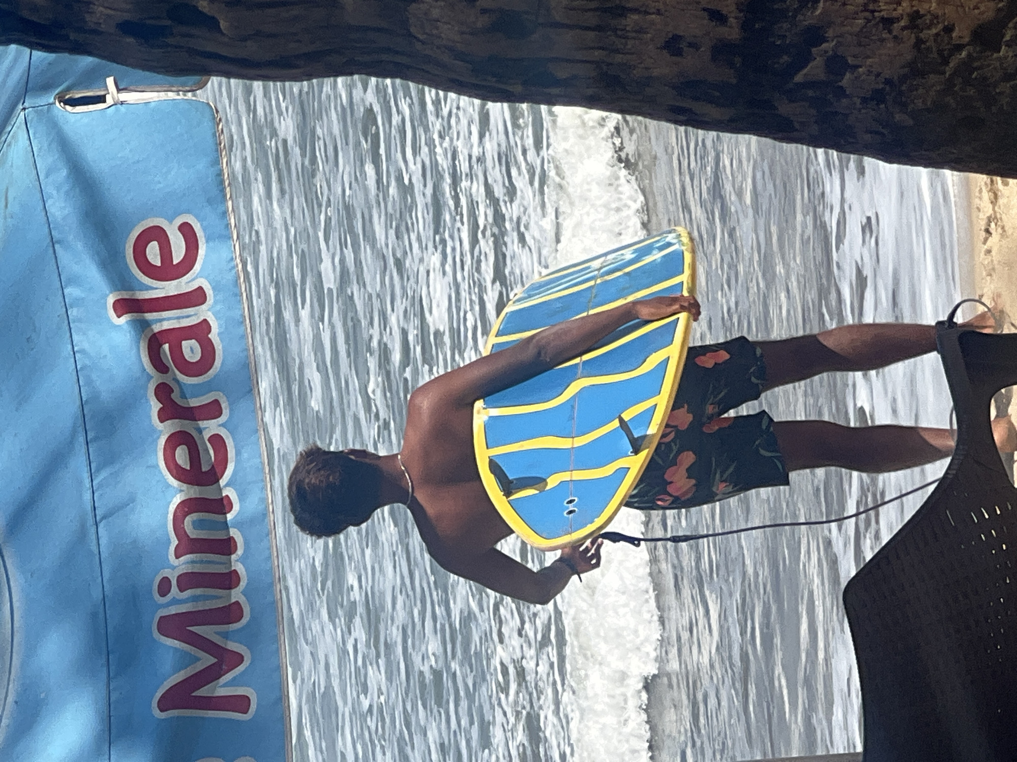 A young man, viewed from behind, stands on the sandy shore of Kuta Beach, Indonesia, holding a blue and yellow boogie board. He is wearing dark patterned swim trunks. In the background, gentle waves roll in. Above the beachgoer, a blue canopy with red lettering that reads "Minerale" is visible, suggesting a nearby refreshment stand or a branded umbrella. The scene is framed by the dark bark of a palm tree on the right side of the image. The lighting indicates it is daytime, with the sun casting shadows.