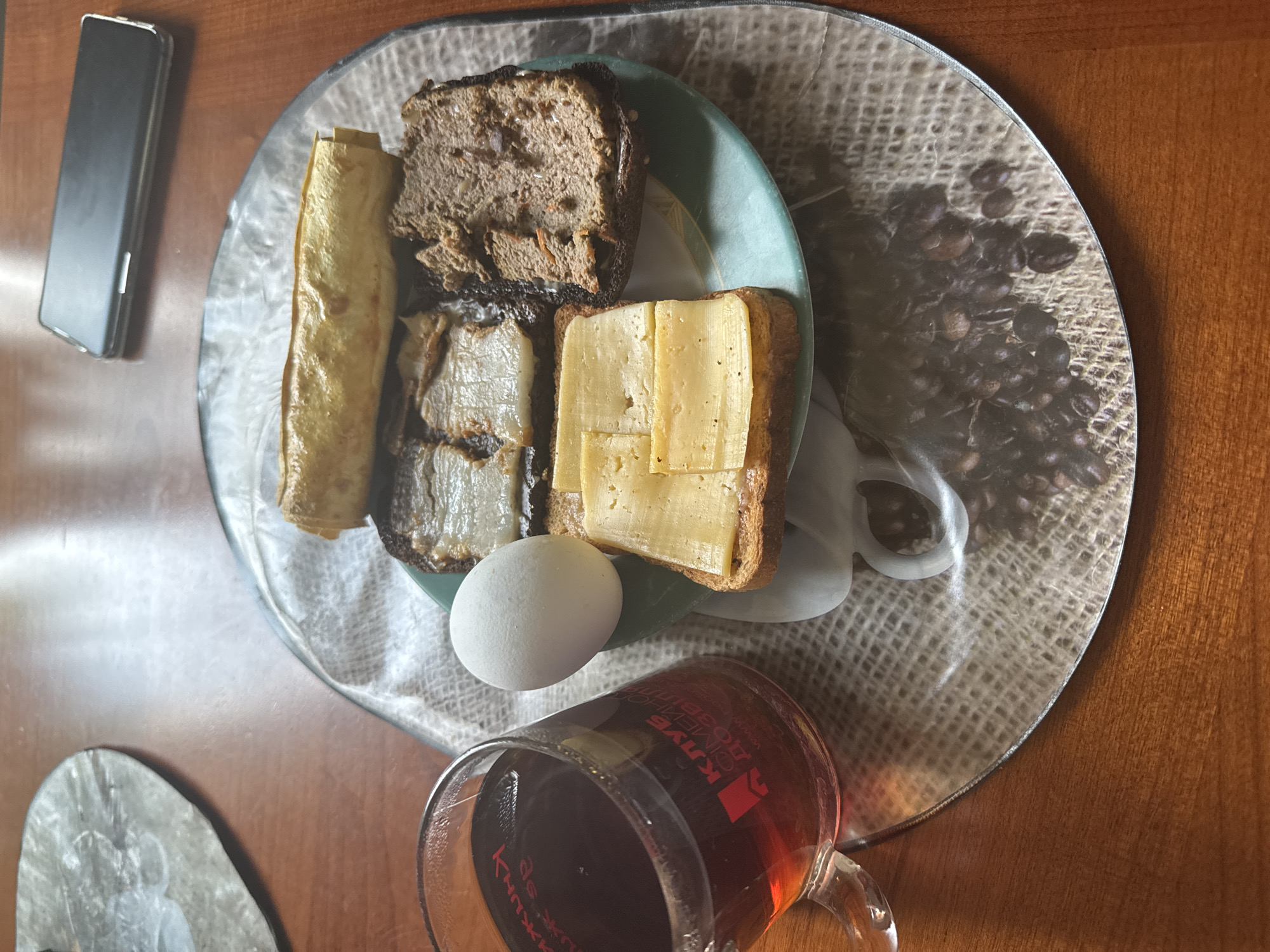 This image, taken in Odesa, Ukraine, depicts a meal set on a reddish-brown wooden table. A clear, circular placemat with a printed design resembling burlap and scattered coffee beans supports a light teal plate. On the plate, there are four distinct food items: a slice of toasted bread topped with multiple rectangular slices of light yellow cheese; a slice of dark bread spread with a brown, textured substance resembling pate; two slices of dark bread topped with pieces of white, semi-translucent rendered fat or lard; and a single golden-brown rolled item, likely a crepe or flatbread. Adjacent to the plate, a whole, white, boiled egg is present. To the left, a clear glass mug containing dark brown tea displays red Ukrainian text, including "КЛУБ" and "СІМЕЙНОГО ДОЗВІЛЛЯ". A dark-colored smartphone with a silver edge lies face down on the table in the upper right portion of the frame. A second, identical placemat is partially visible in the upper left corner. No individuals are visible in the scene.