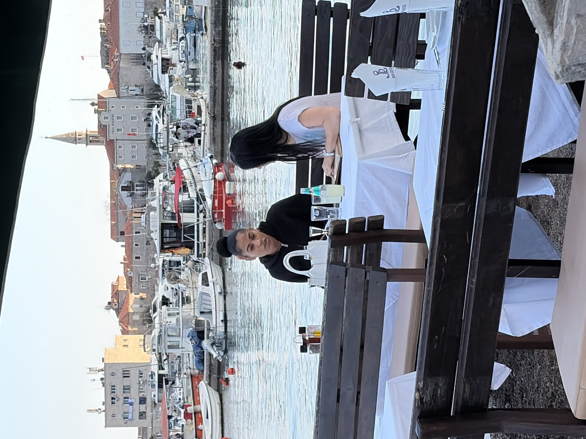 The image captures an outdoor dining scene in Budva, Montenegro. Two women are seated at a table adorned with a white tablecloth. The woman on the left wears a black hooded sweatshirt, while the other woman, seen from the back, has long, dark hair and wears a light-colored top. The table is set with glassware, bottles, and condiments. A white handbag is visible near the woman in the black sweatshirt.
In the background, a harbor is filled with boats, and buildings are visible along the waterfront. The architectural style indicates that it's Budva. The sky appears overcast. The dining area includes dark wooden benches.