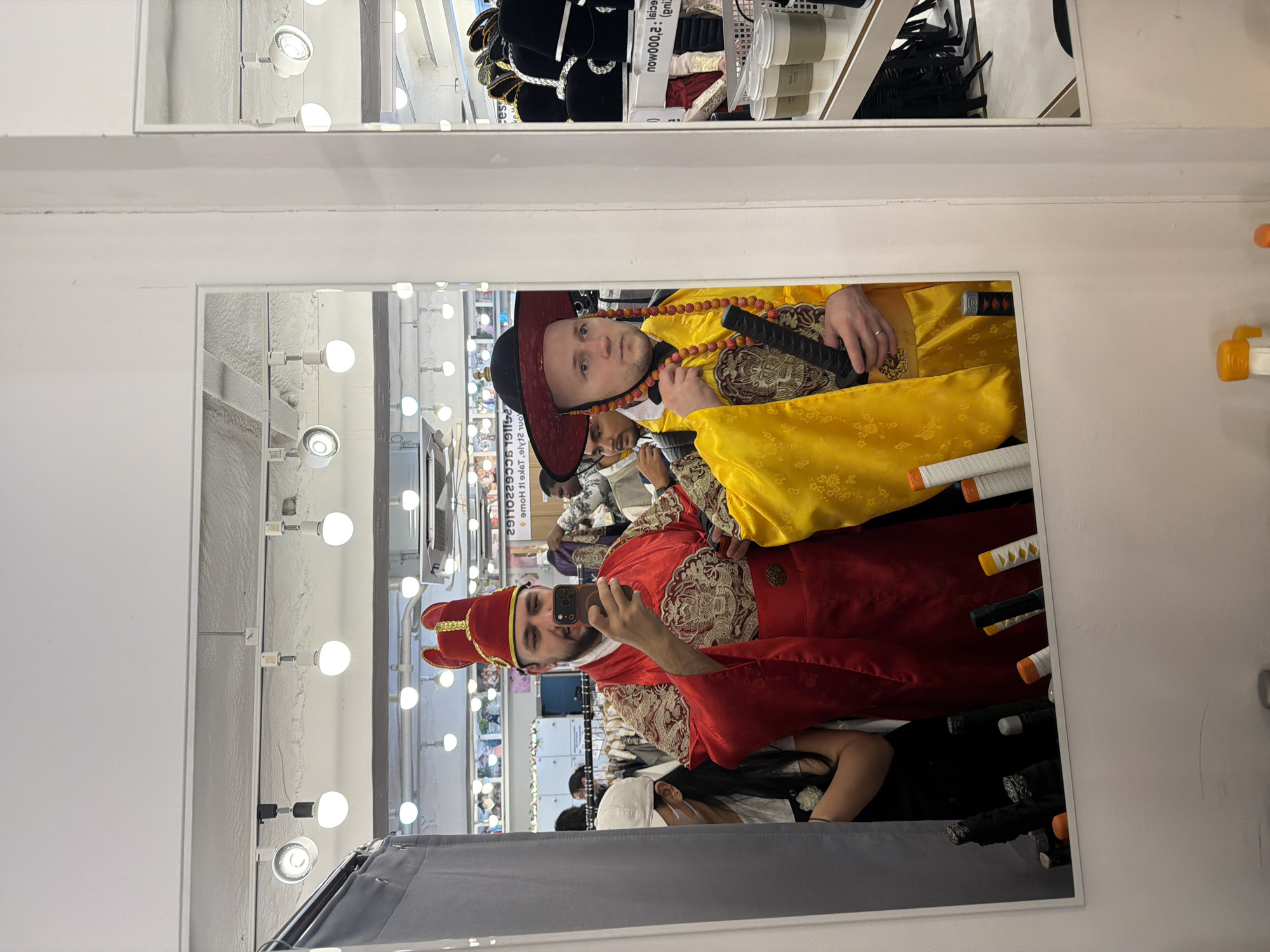 The image depicts a reflection in a large mirror within a traditional costume rental or retail store in Seoul, South Korea. Two adult males are prominently featured.
The male on the left, with a beard, wears a red traditional Korean-style costume (hanbok) featuring elaborate gold embroidery on the chest and sleeves, and a tall, red, winged hat with gold trim. He holds a black smartphone in his right hand, pointed towards the mirror, indicating he is taking a photograph or video. A female individual, partially visible behind him on the left, wears a white baseball cap and a white face mask, with a dark top.
The male on the right, bald, wears a vibrant yellow traditional Korean-style costume with similar gold embroidery and a wide-brimmed black hat with red trim, adorned with dangling red and orange beaded strands. He holds a black, sheathed object resembling a sword or staff in his right hand. His gaze is directed slightly to the viewer's right. Behind him, another male figure is partially visible.
The background, reflected in the mirror, shows a retail environment with multiple overhead track lights and round bulbs illuminating white walls and ceiling. Racks displaying various garments and shelves stocked with hats, including black tall hats with silver accents, are visible. Below the mirror, several prop weapons or decorative items, including stylized swords with orange and white hilts, are arranged. To the far right, a shelf holds several disposable beverage cups. The reflected text on a sign indicates "SPECIAL: 2,000WON," confirming the South Korean location.