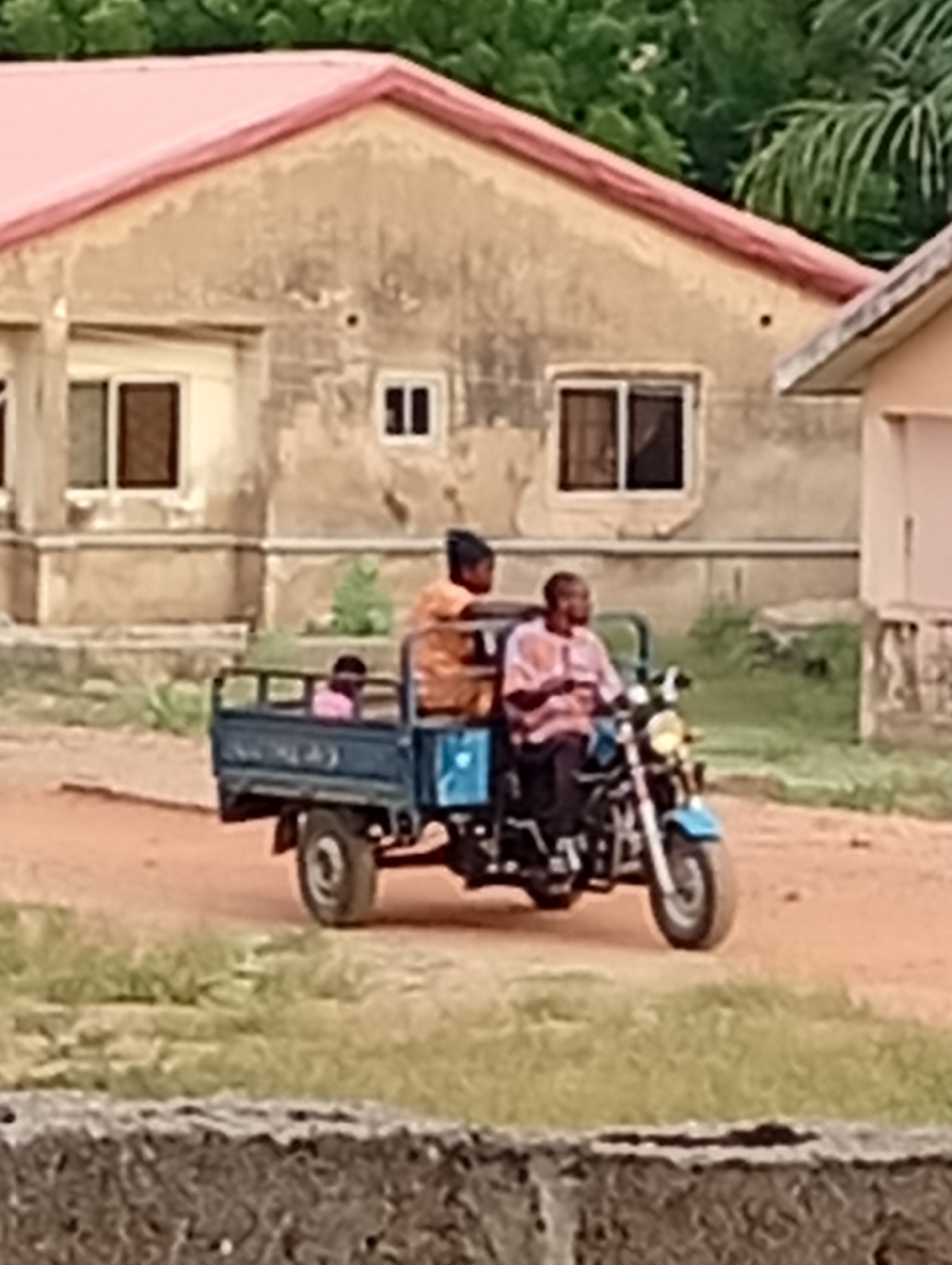 The image shows a motorized tricycle on a dirt road in front of two buildings with pink roofs. The main building has three windows visible, while the second building is partially visible on the right. A man is driving the tricycle, and two passengers are in the cargo area, one appearing to be a child. The area appears to be in a city, but the exact city and location are Unknown, Unknown. Vegetation is visible in the background.