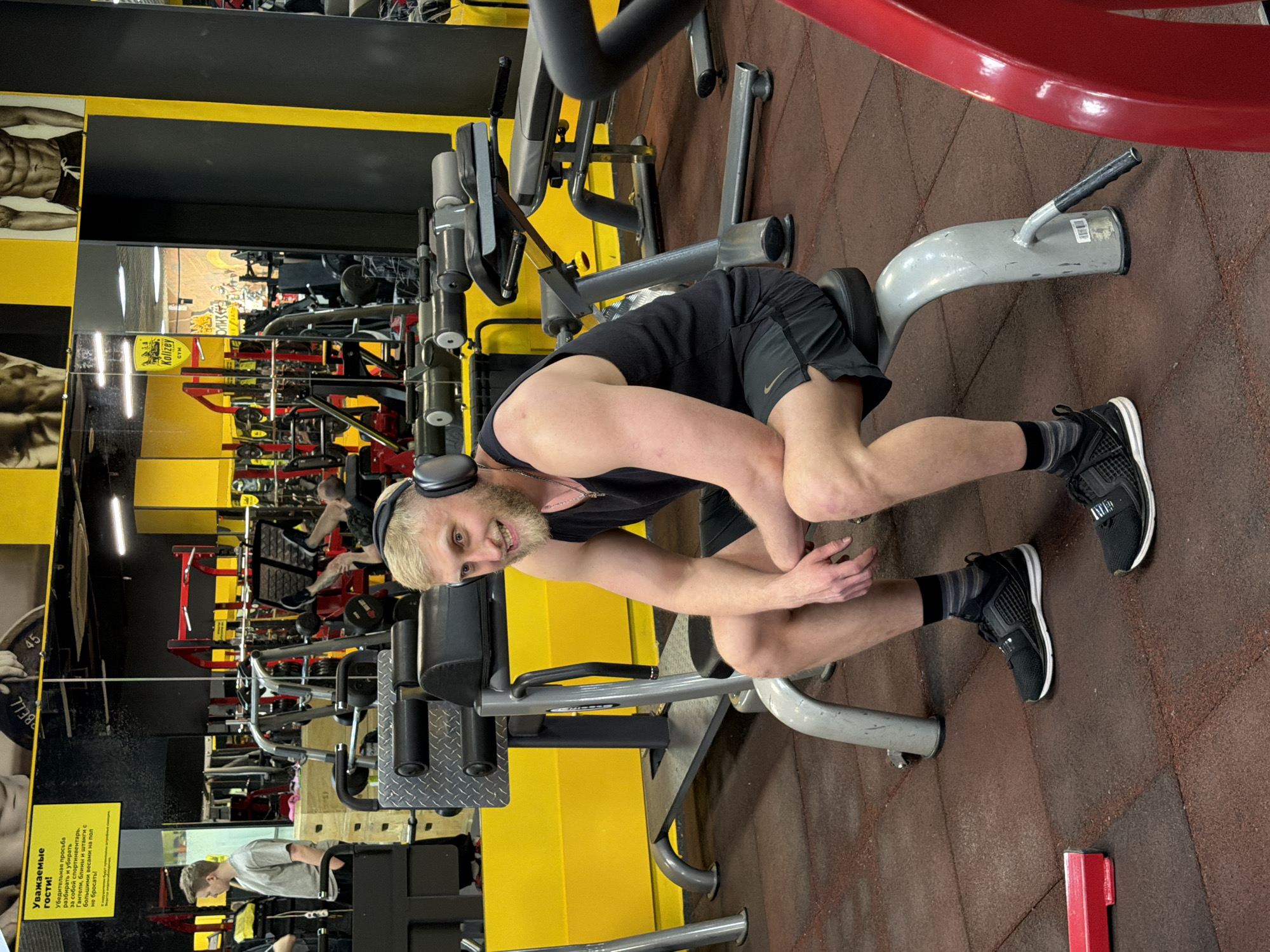 A man with blond hair and a beard is in a gym in Krasnoyarsk, Russia. He is wearing a black tank top, black shorts, and black sneakers with white soles and is positioned on a piece of exercise equipment. He has headphones on his head. The gym has various exercise machines, and the floor is a reddish-brown color. A sign in Russian is visible on the wall, and there are posters of bodybuilders.