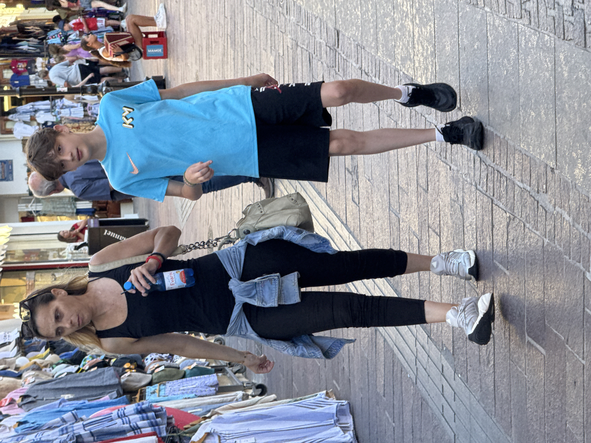 In Chania, Greece, a woman and a boy walk down a cobblestone street lined with shops. The woman, with blonde hair pulled back and wearing sunglasses on her head, is dressed in a black tank top, black leggings, and gray sneakers. She holds a blue bottle of Evian water in her right hand and has a light-colored shoulder bag slung across her body. A light blue denim jacket is tied around her waist. The boy, with short brown hair, wears a bright blue t-shirt with a KY logo and black athletic shorts with white and red accents. He is also wearing black sneakers and white socks. The street is busy with other pedestrians, and storefronts with clothing displays are visible on either side. In the background, a person sits playing an accordion.