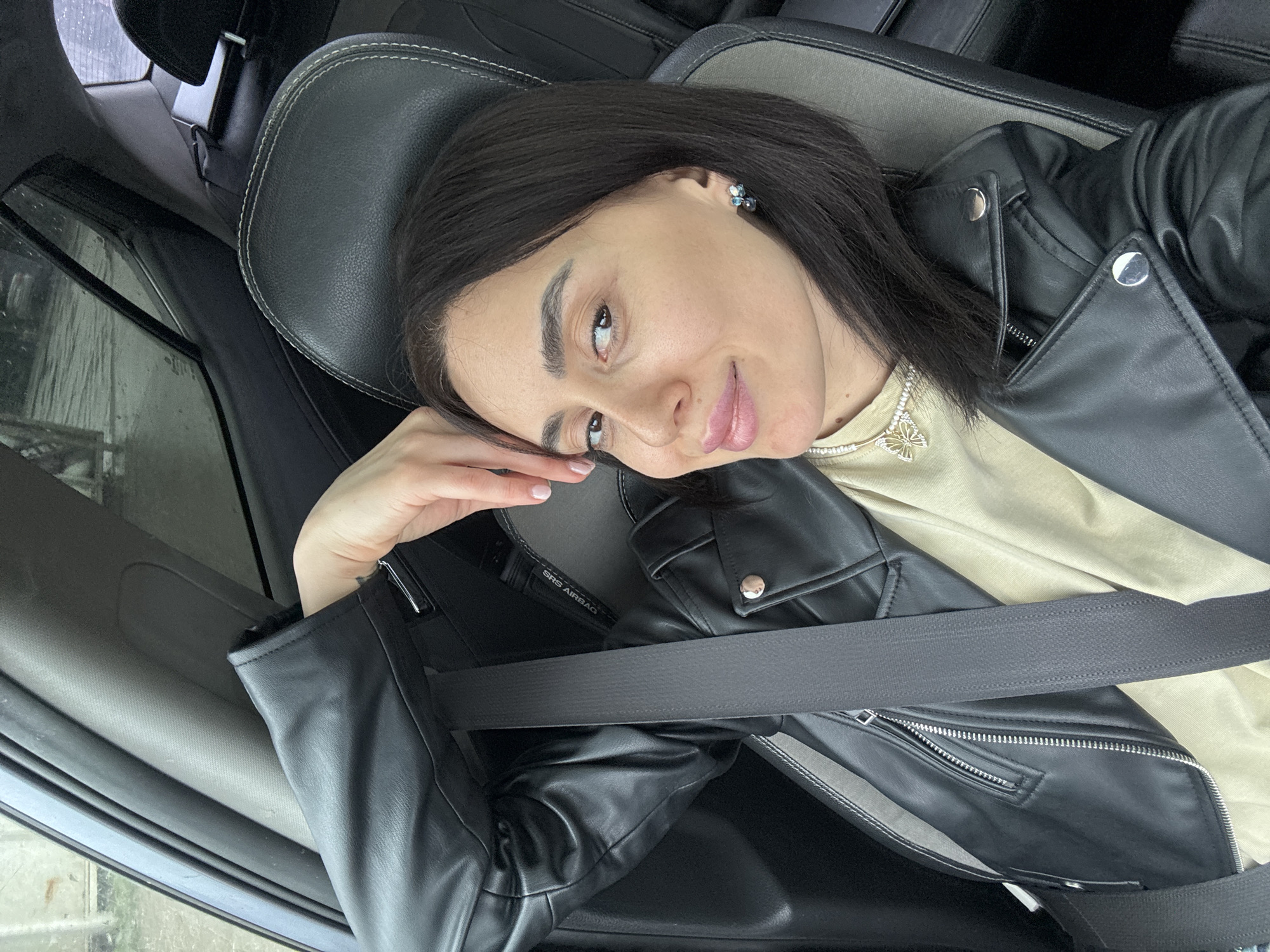 A female individual is depicted in the front passenger seat of a vehicle, angled towards the camera. She is wearing a black faux leather jacket over a light beige top, with a black seatbelt secured across her chest and lap. A necklace with a butterfly-shaped pendant is visible around her neck, and a blue and silver cluster earring is present on her right ear. Her right hand is positioned with fingers extended, resting on the upper rear section of the car seat behind her head. The car interior features black leather or faux leather seats with light grey stitching, and dark grey trim elements. An "SRS AIRBAG" label is visible on the vehicle's B-pillar. Through the windows, an exterior environment is visible, characterized by wet ground and indistinct architectural elements, suggesting an urban or suburban area under damp, overcast conditions. The specific location within Tbilisi, Georgia, cannot be determined from the visual information provided.