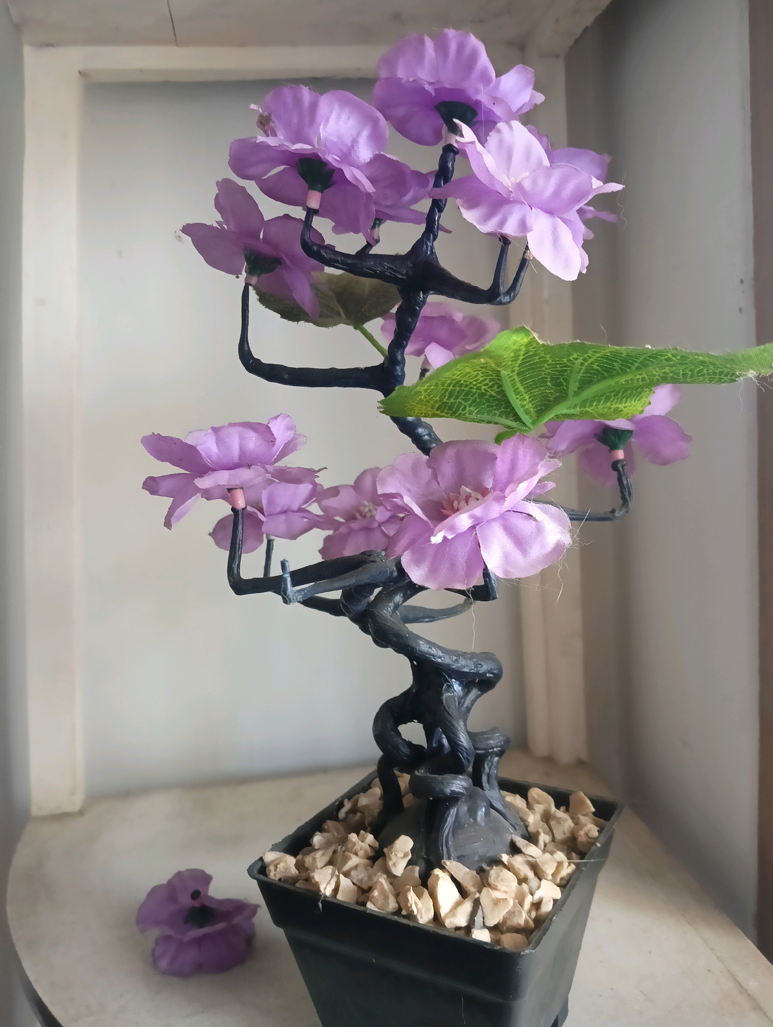 The image shows a decorative artificial plant with purple flowers and a green leaf, placed in a black pot filled with small stones. The plant has a twisted black trunk and branches. To the left of the pot, a single fallen purple flower lies on a white surface. The background consists of a white framed area and a beige wall. The scene is indoors, likely on a shelf or windowsill. The specific location within Kromengan, Indonesia, cannot be determined from the image alone.
