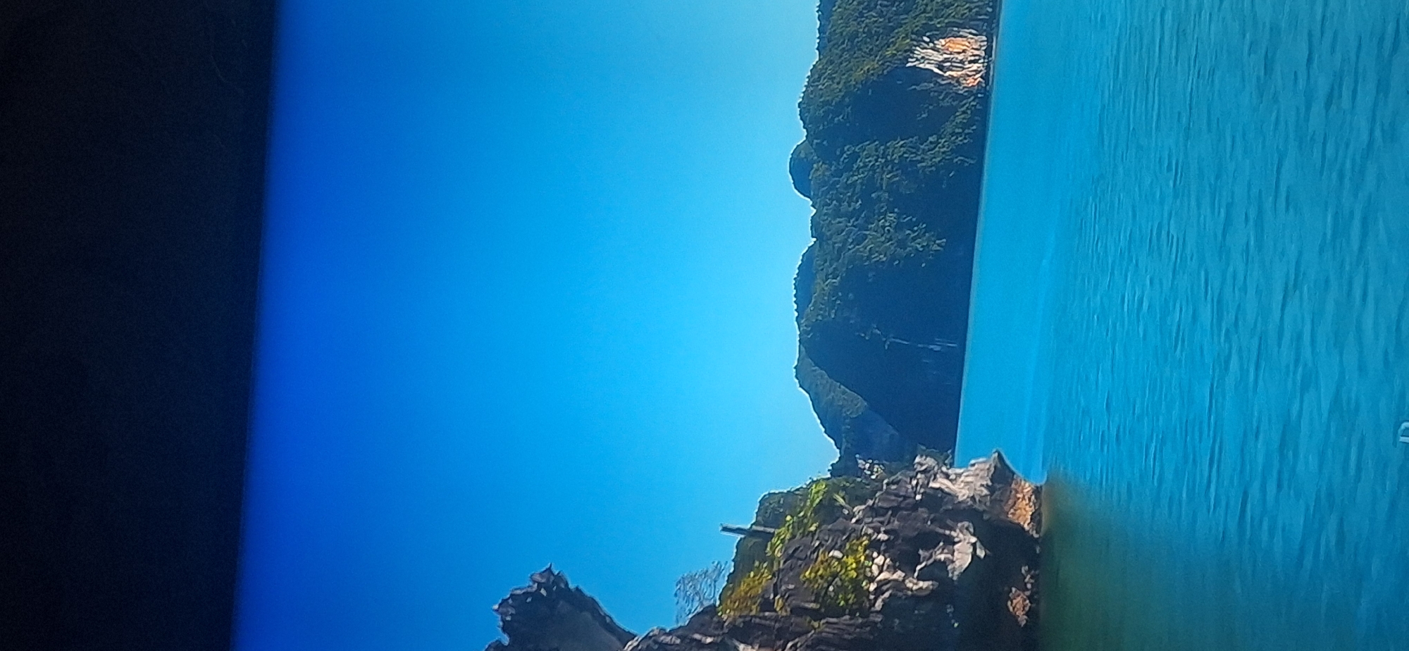 The image depicts a scenic view of a body of water, likely a bay or sea, with numerous verdant, tree-covered karst islands and limestone formations rising from the water. The water is a vibrant turquoise blue, with gentle ripples on its surface. The sky above is a clear, deep blue, suggesting a sunny day. On the left side of the image, a large, rugged rock formation juts out into the water, its surface textured and weathered. This formation appears to be covered in sparse vegetation in places. In the background, a chain of larger islands, also densely forested, stretches across the horizon. One of these islands shows a distinct, lighter-colored cliff face with an orangish hue. The overall scene evokes a sense of natural beauty and tranquility. The image does not contain any people, objects, or specific details that would place it within the city of Bila Tserkva, Ukraine.